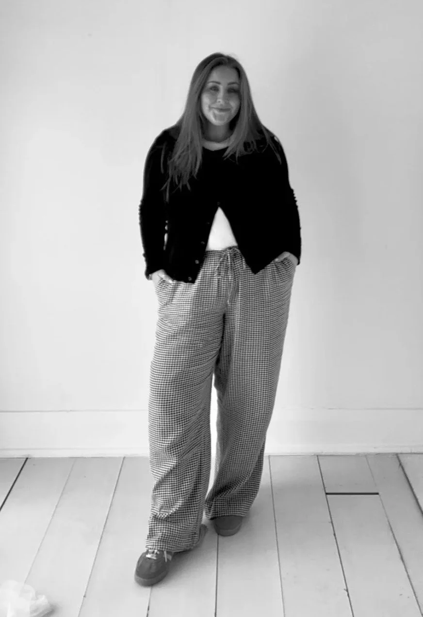 Black and white photo of a woman standing with hands in pockets, smiling, wearing a dark cardigan, light-colored wide-leg checkered pants, and sneakers, in front of a plain wall and wooden floor.