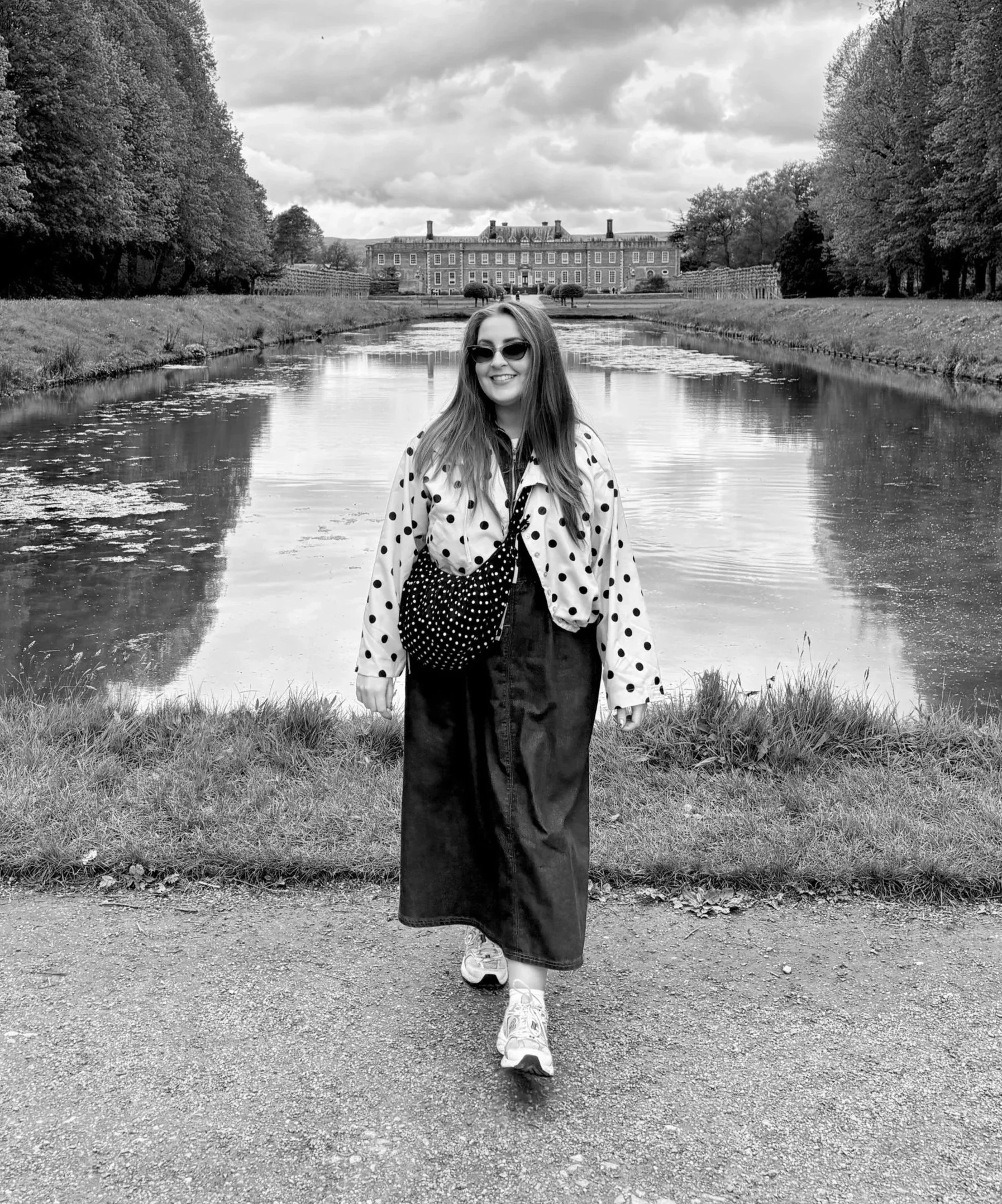 A woman wearing sunglasses, a polka dot jacket, and sneakers walking down a gravel path beside a calm river, with a large historic building and trees in the background.