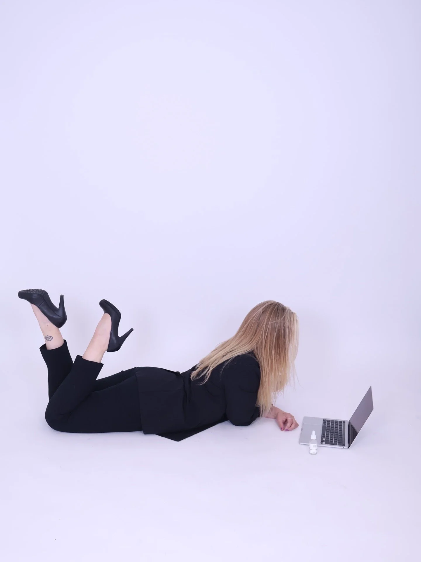 Woman lying on her stomach on a white background, working on a laptop, with her legs bent at the knees and crossed at the ankles, wearing black high heels and black clothing, with a small bottle and a digital device beside her.