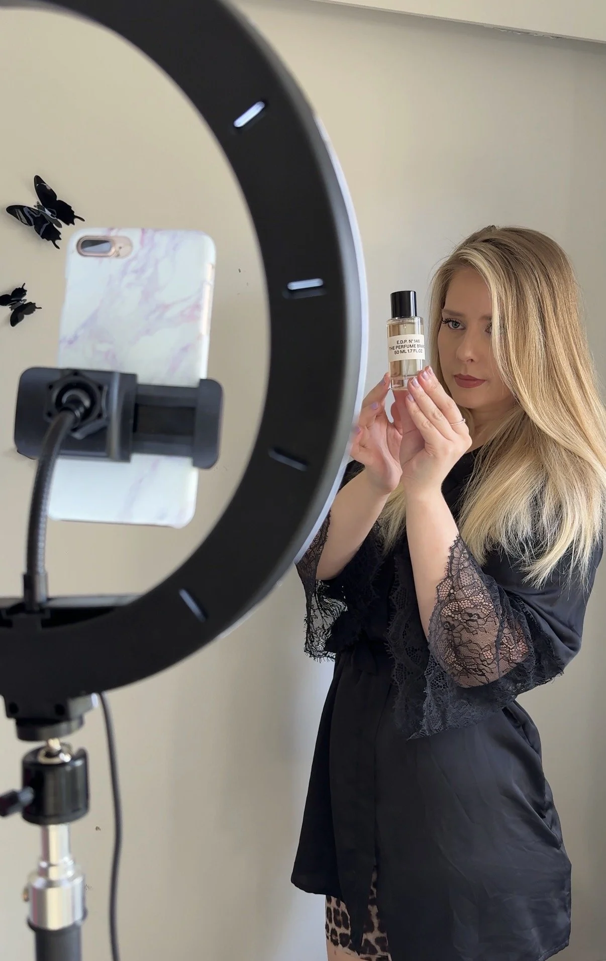 A woman with long blonde hair holding a perfume bottle in front of a mirror, with a ring on her finger, standing against a beige wall with black butterfly decorations.