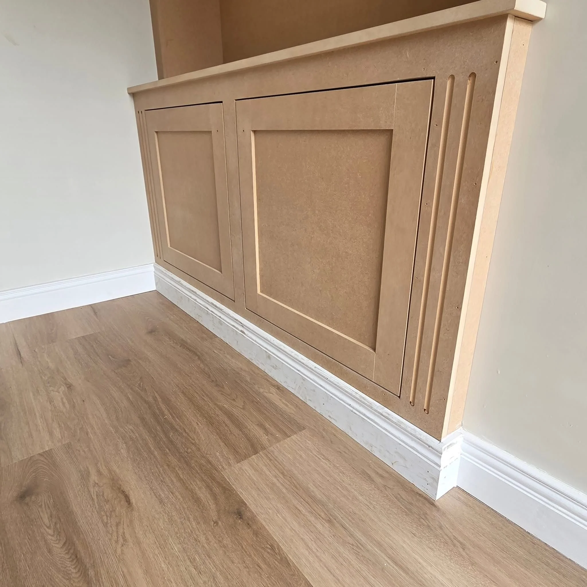 A wooden fireplace mantel with decorative molding, set against a wall with light-colored paint and white crown molding. The floor beneath is hardwood.
