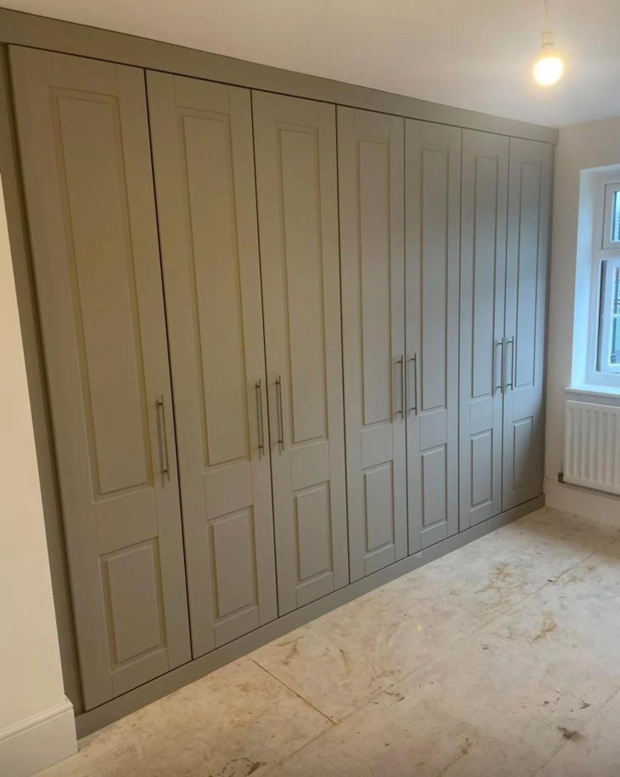 A row of beige kitchen cabinets with metal handles, positioned along a wall in a room with a window and a light fixture.
