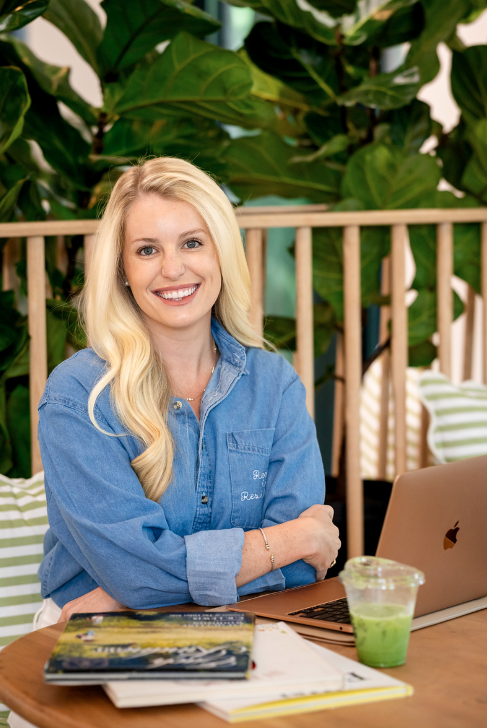 A young woman with long blonde hair wearing a blue denim shirt, smiling and sitting at a table with a laptop, a green drink, and some books, in a setting with lush green plants.