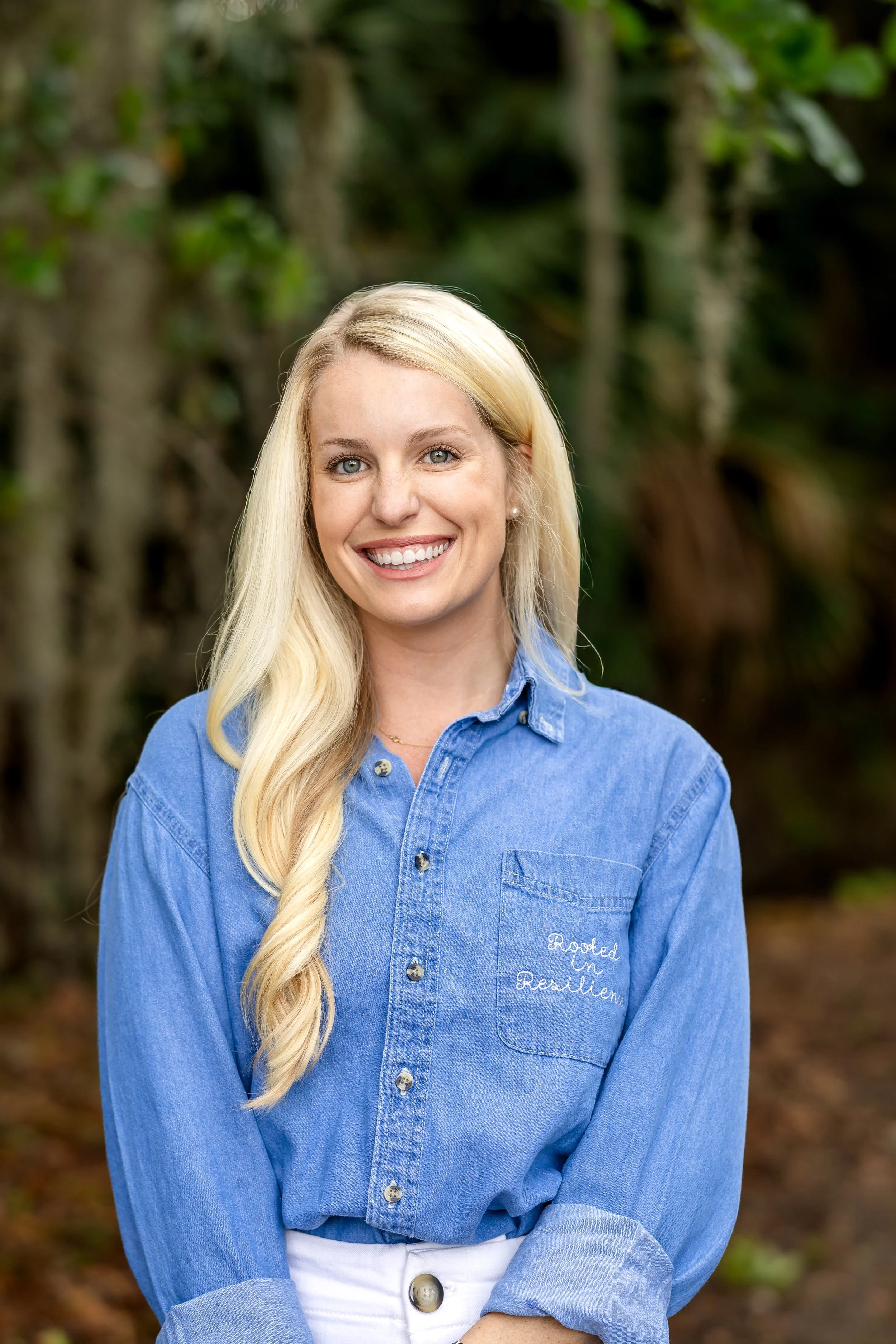 A smiling woman with long blonde hair wearing a blue shirt with embroidered text, standing outdoors with a blurred green background of trees and foliage.