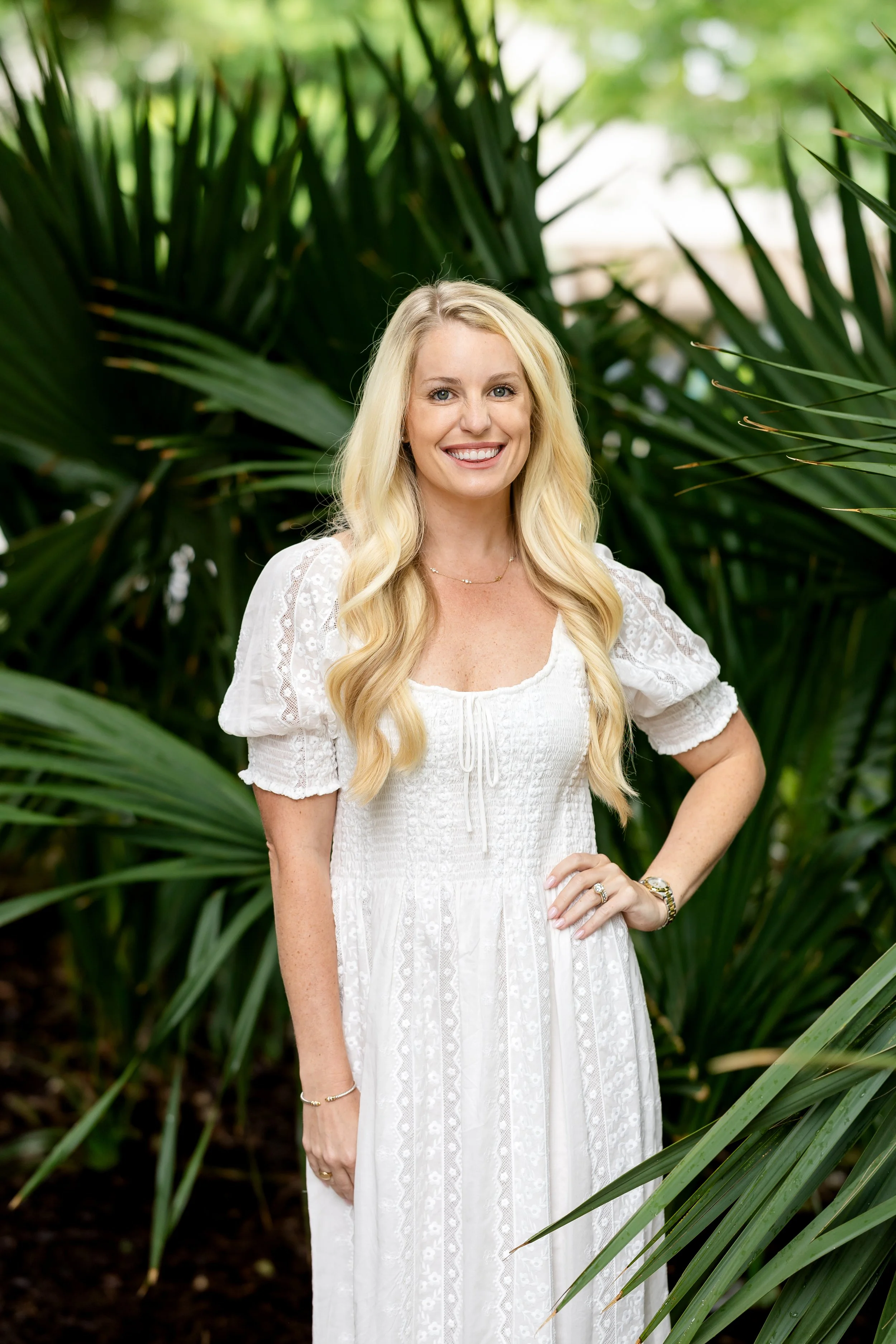 A woman with long blonde hair wearing a white dress, standing outdoors among green plants, smiling at the camera.