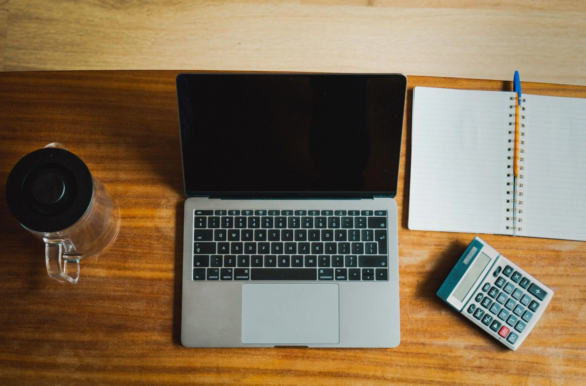 Top-down view of a wooden desk with a laptop, a spiral notebook with a blue pen, a calculator, a water bottle, and a small box.