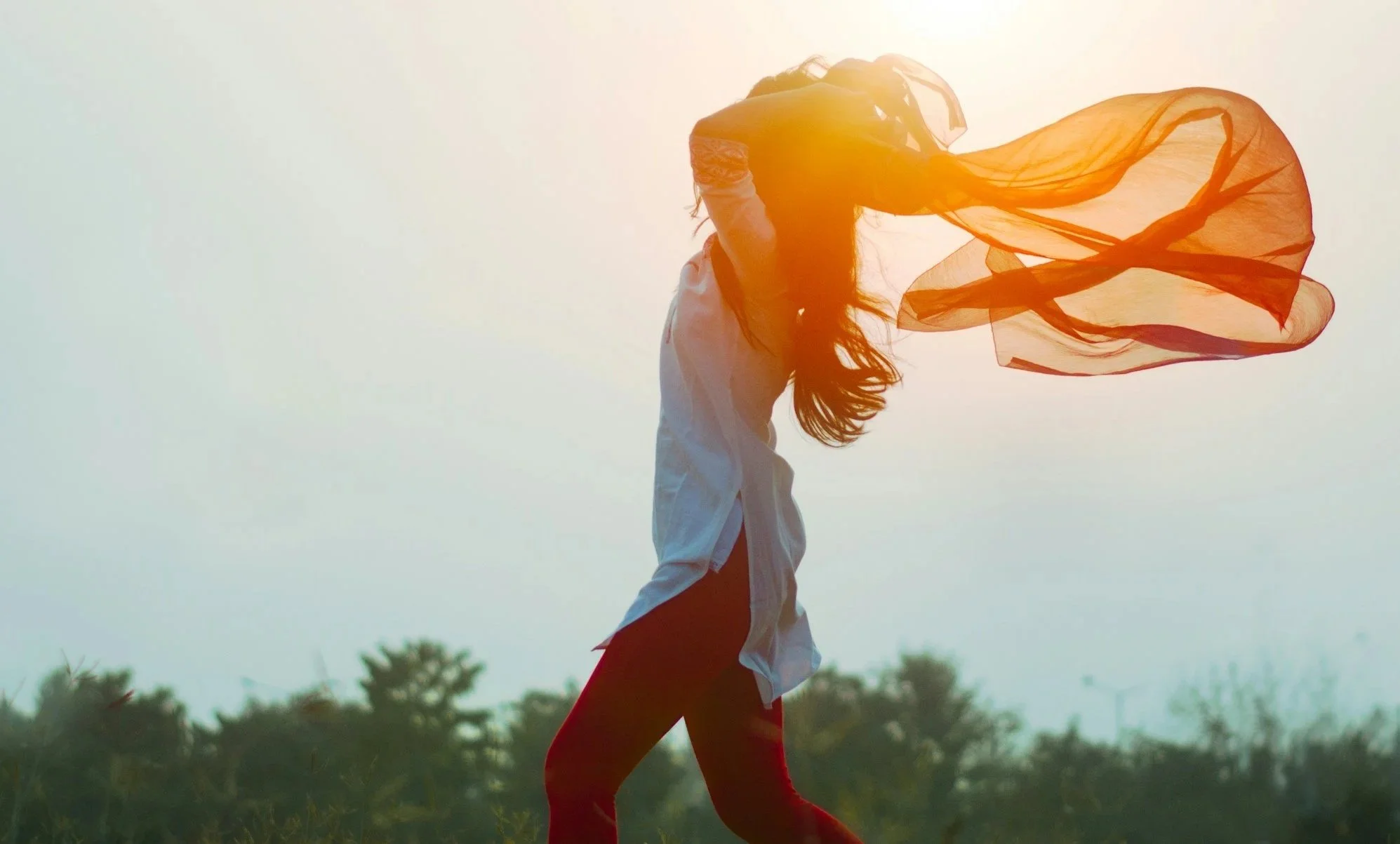 A woman outdoors holding a large orange scarf with sunlight in the background.