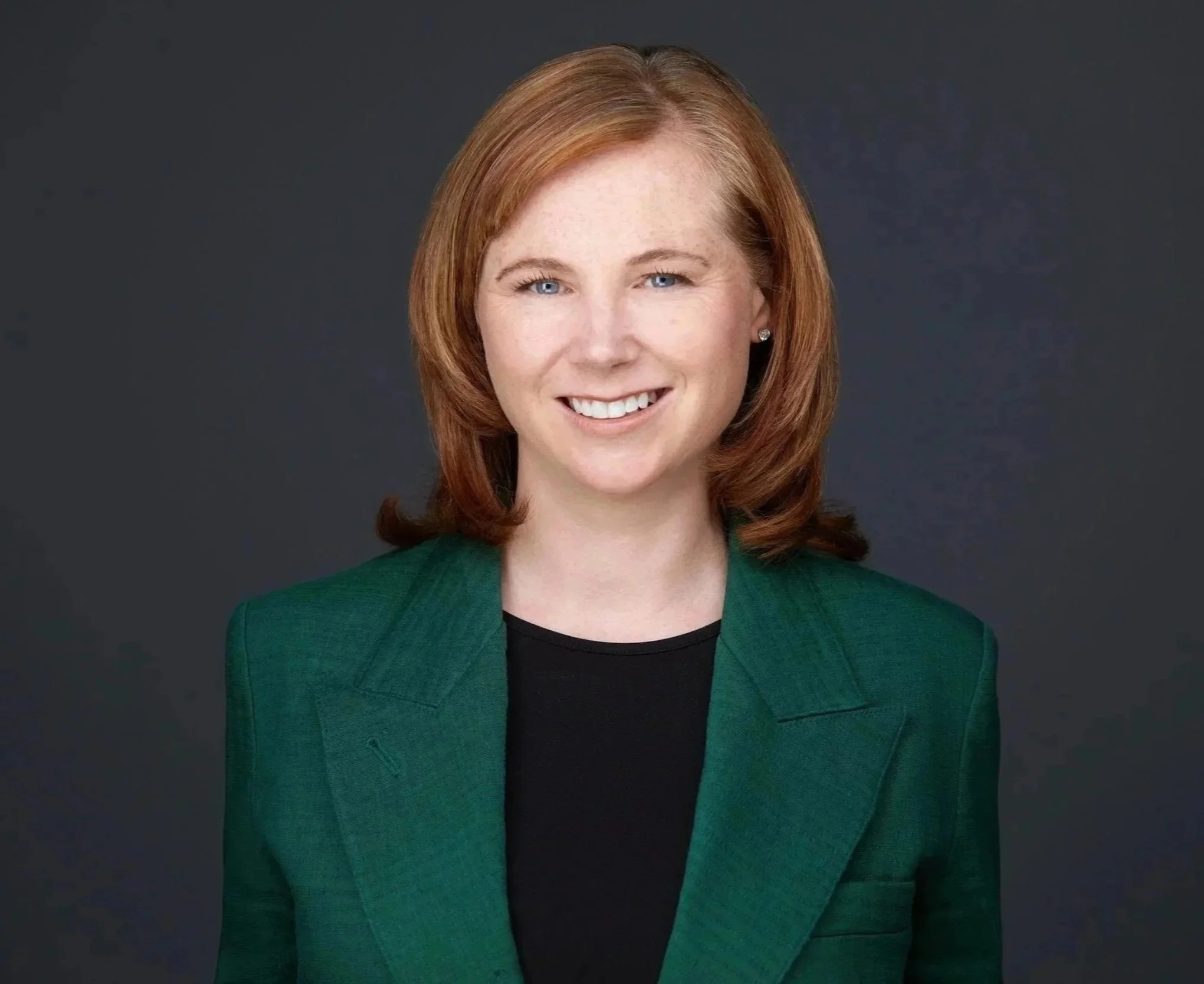 A headshot of a smiling woman with red hair, wearing a green blazer and black top, against a dark background.