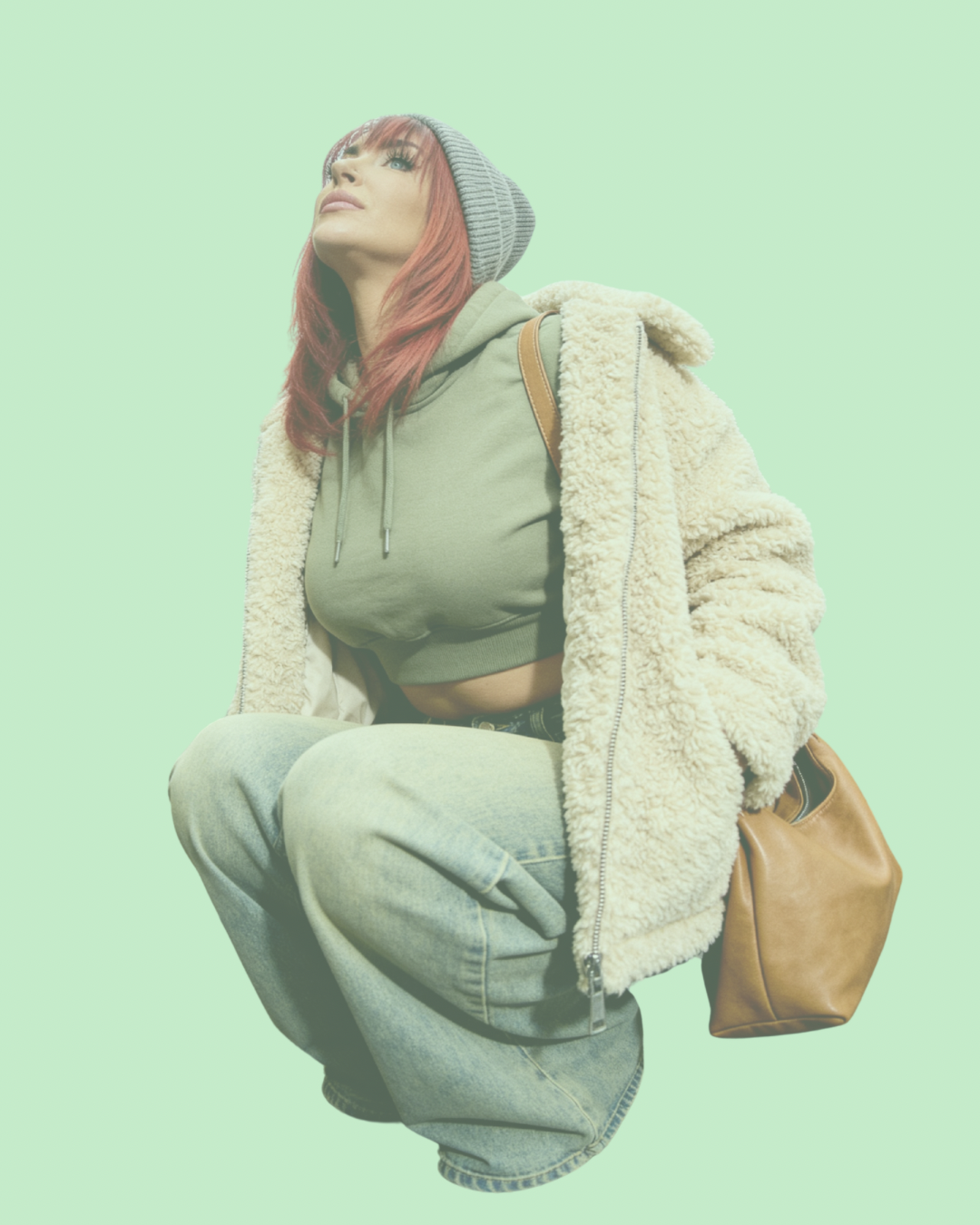 A young woman with red hair, wearing a gray beanie, olive green hoodie, beige shearling jacket, and light-wash jeans, squatting with a brown backpack against a light green background.