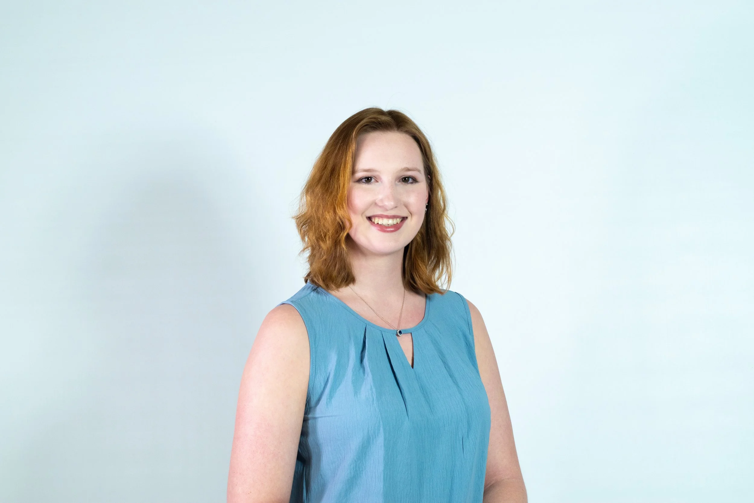 A young woman with red hair, blue sleeveless top, smiling, against a plain light blue background.