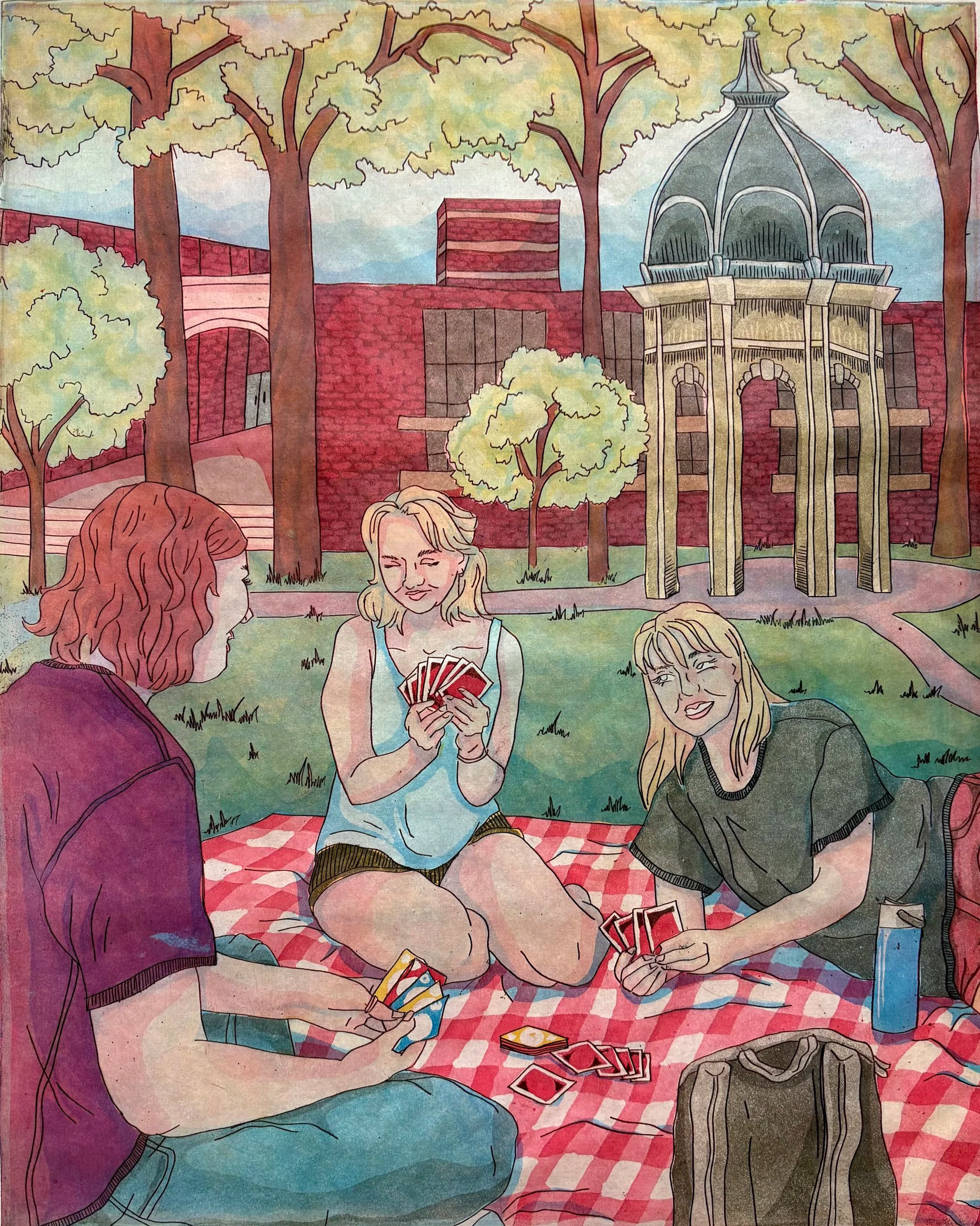 Three young women playing cards outdoors in a park with trees and a gazebo, on a red and white checkered blanket.