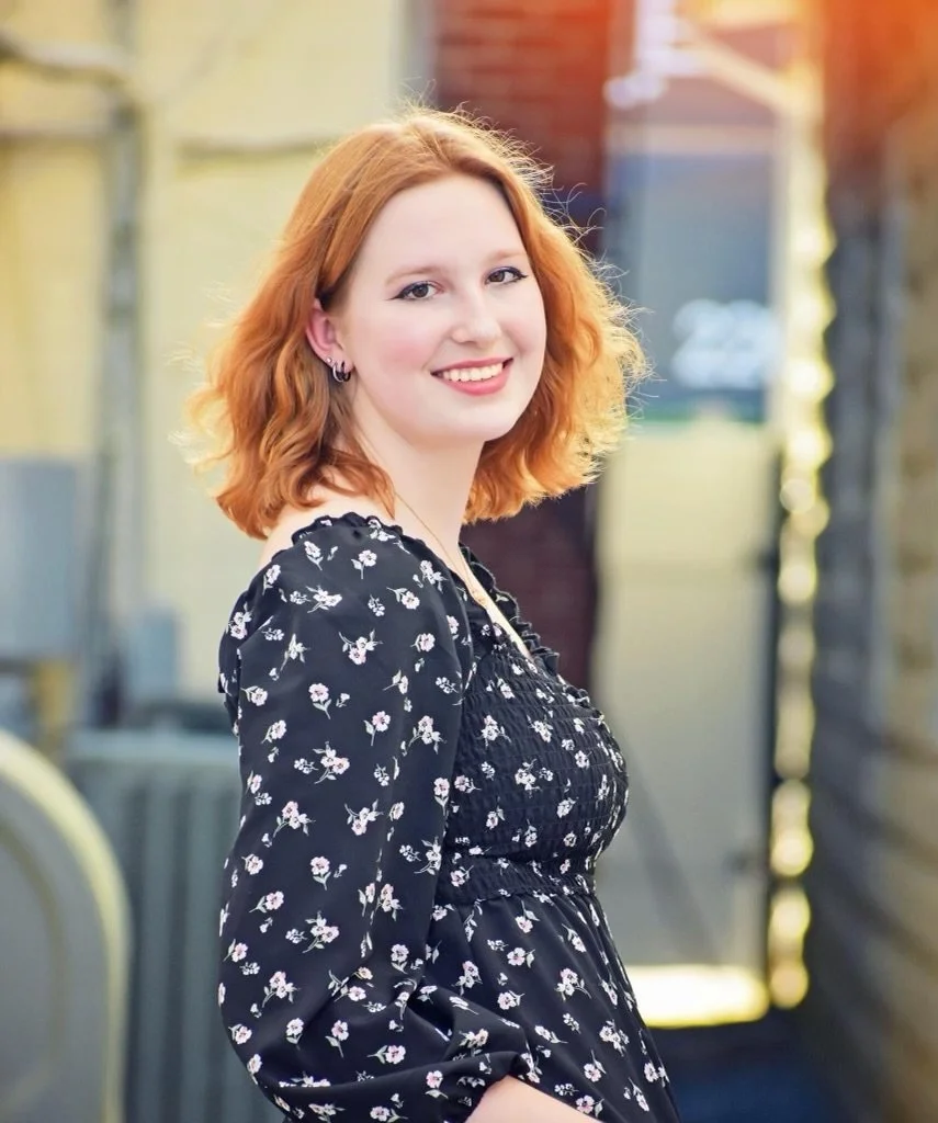 A young woman with shoulder-length red hair, wearing a black floral dress, smiling outdoors in sunlight.