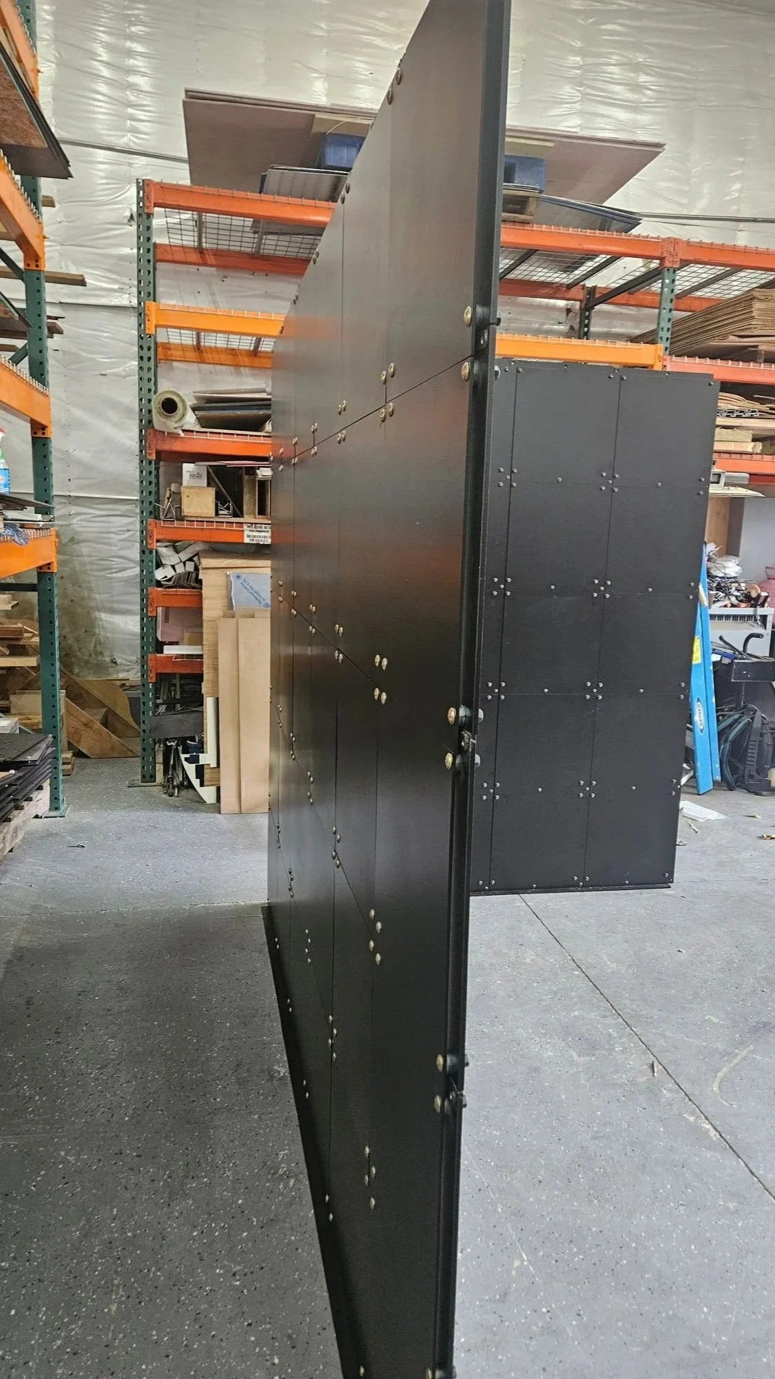 A black panel standing in a warehouse or workshop with orange metal shelves holding various materials and tools in the background.