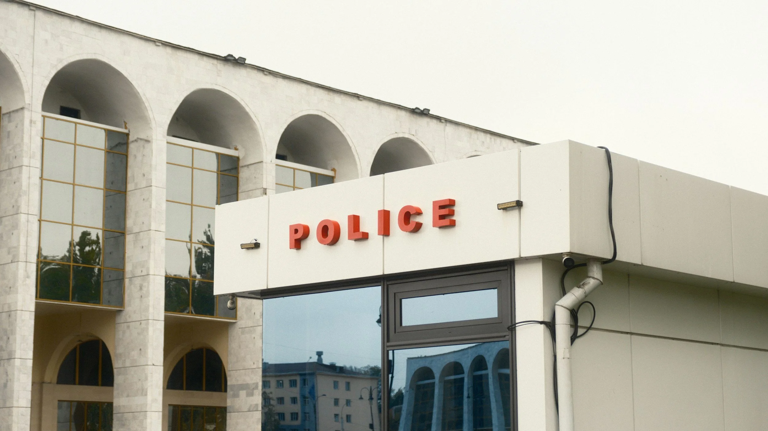 Exterior of a building with a white wall displaying red letters spelling "POLICE" above a window, with a modern architectural design including arched windows on upper floors and reflective glass windows.