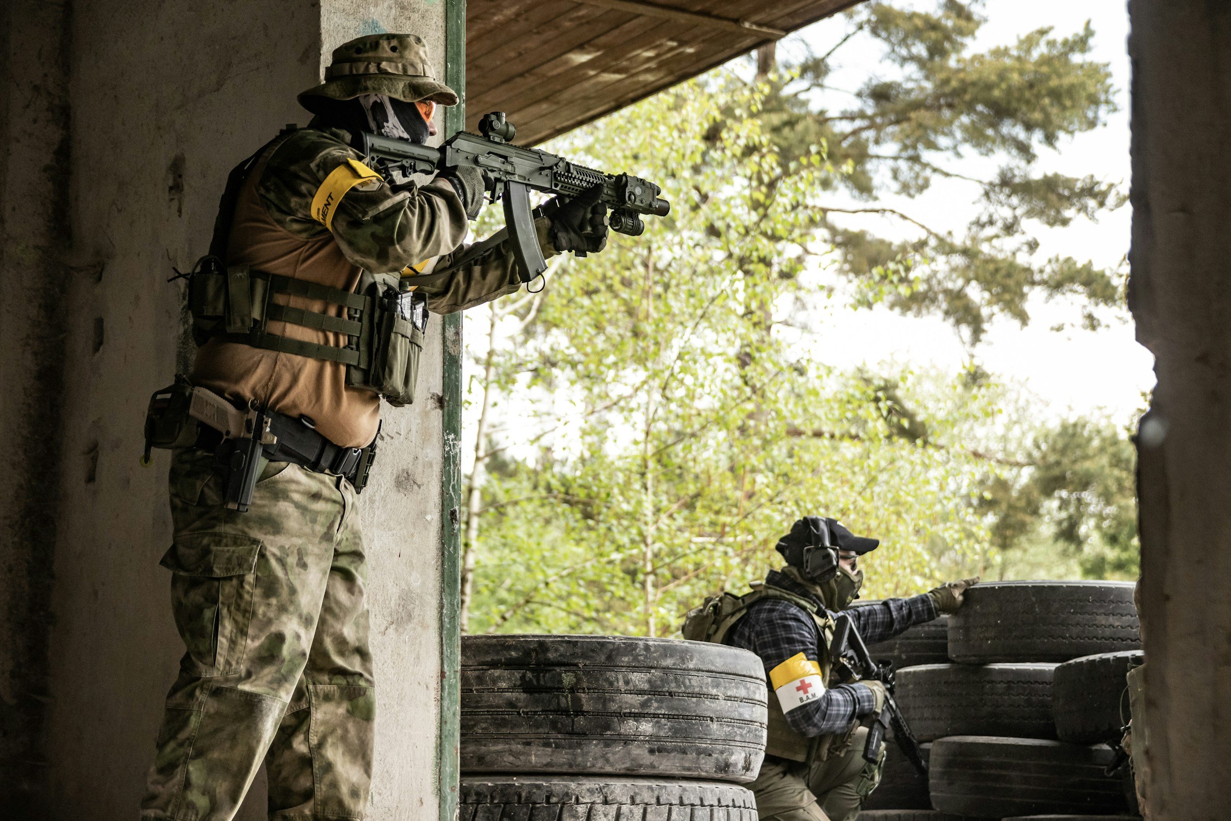 Two soldiers in camouflage uniforms and tactical gear aiming rifles from a fortified position with tires and trees in the background.