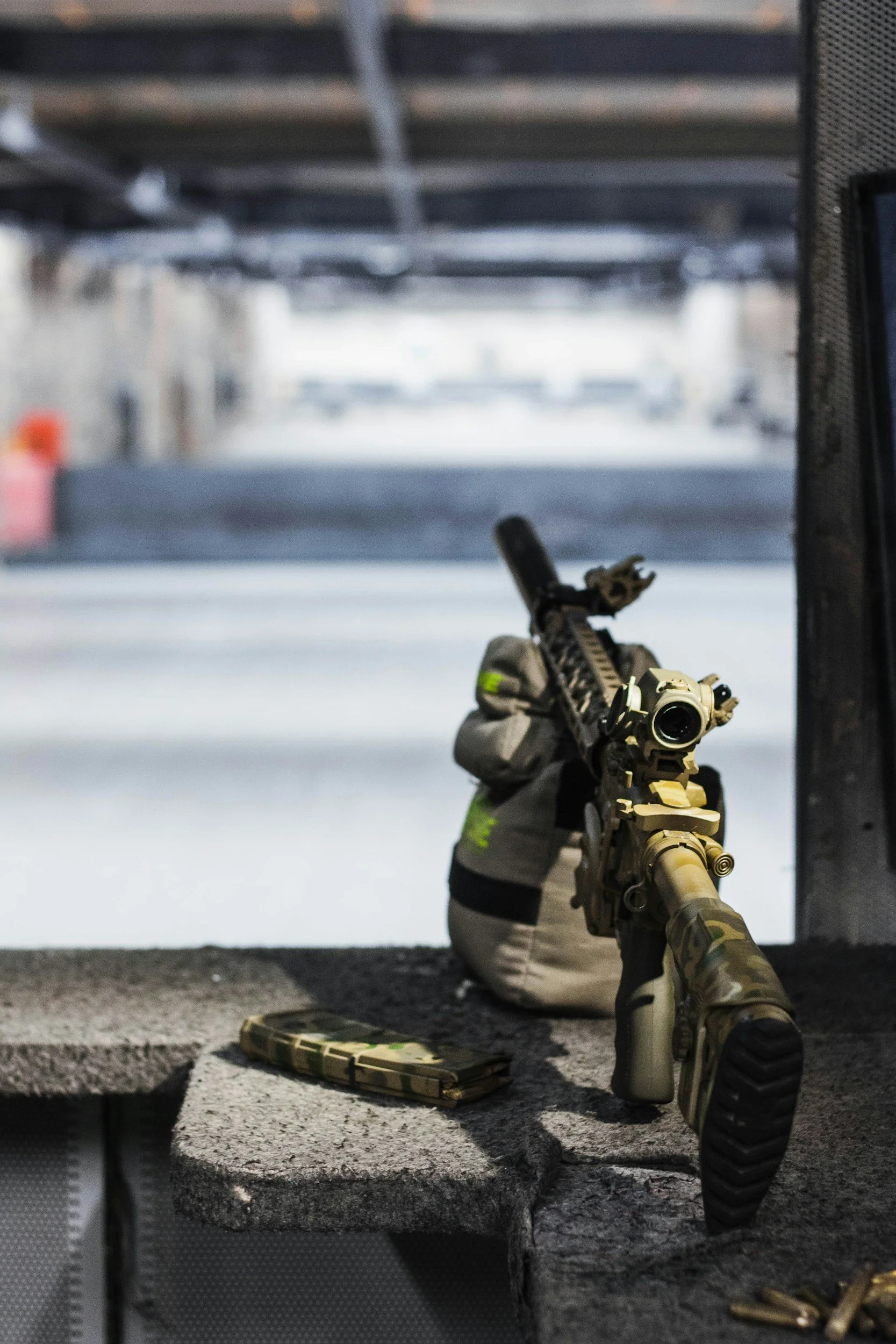 A Tactical rifle with camouflage pattern is placed on concrete steps, with a tan scope mounted on top. A matching camouflage magazine rests on the step in front of it, alongside some brass casings. The background is blurred, showing parts of a parking lot with concrete parking blocks and a building.