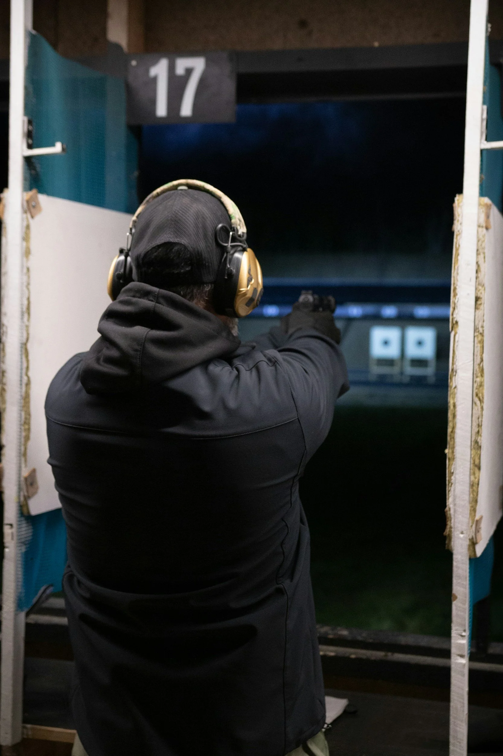 A person in dark clothing and a black beanie, wearing hearing protection, aiming a handgun at an indoor shooting range with targets in the distance. The number '17' is displayed above the shooting area.