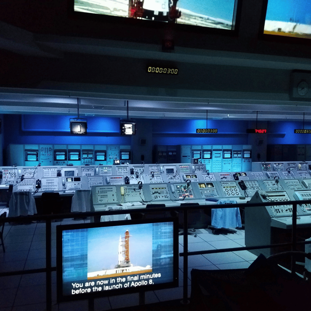 A control room with multiple monitors and electronic equipment illuminated by blue lighting, with a digital countdown timer on the ceiling, and a screen showing a lunar launch scene and the text about the final minutes before Apollo 8 launch.