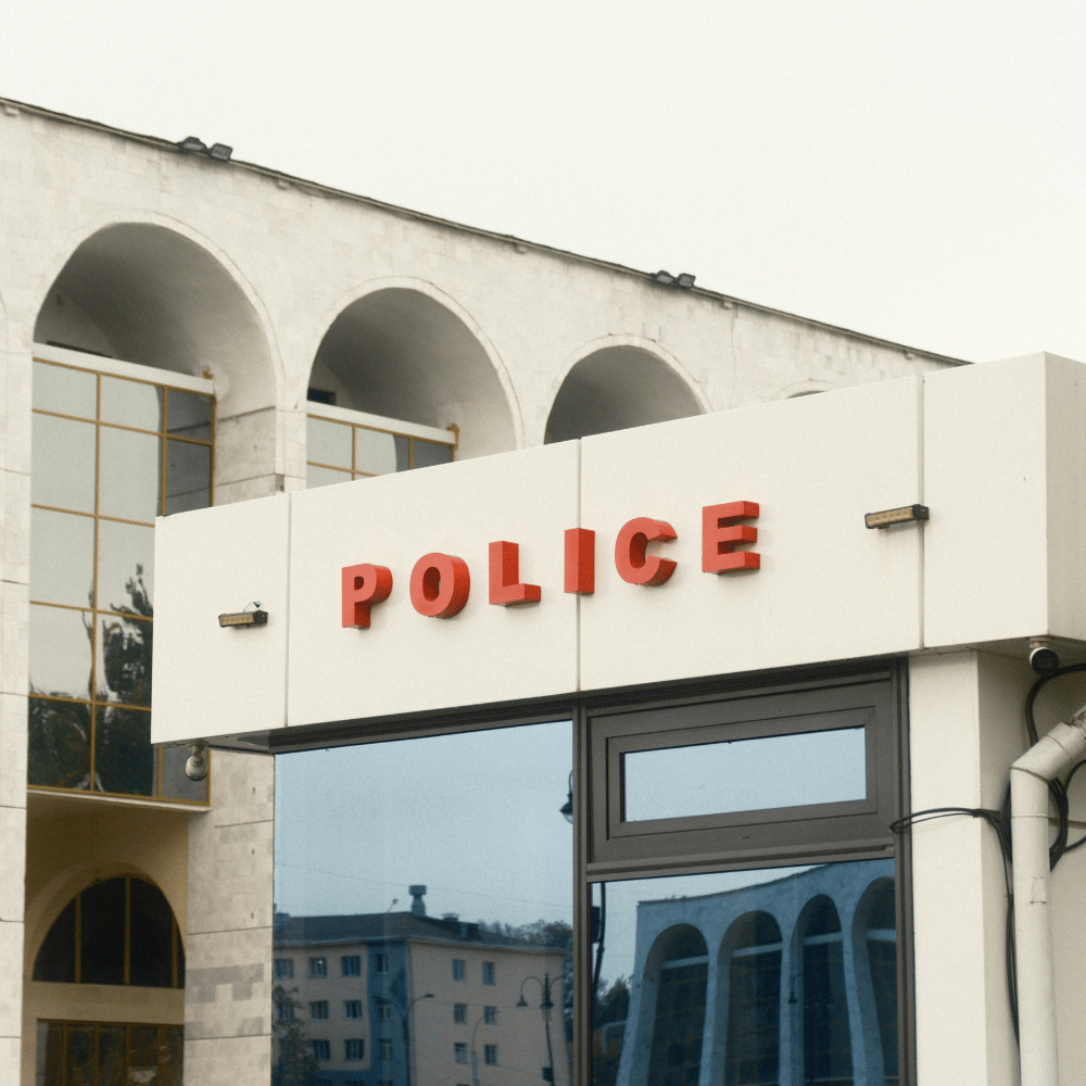 Exterior of a building with a white wall displaying red letters spelling "POLICE" above a window, with a modern architectural design including arched windows on upper floors and reflective glass windows.
