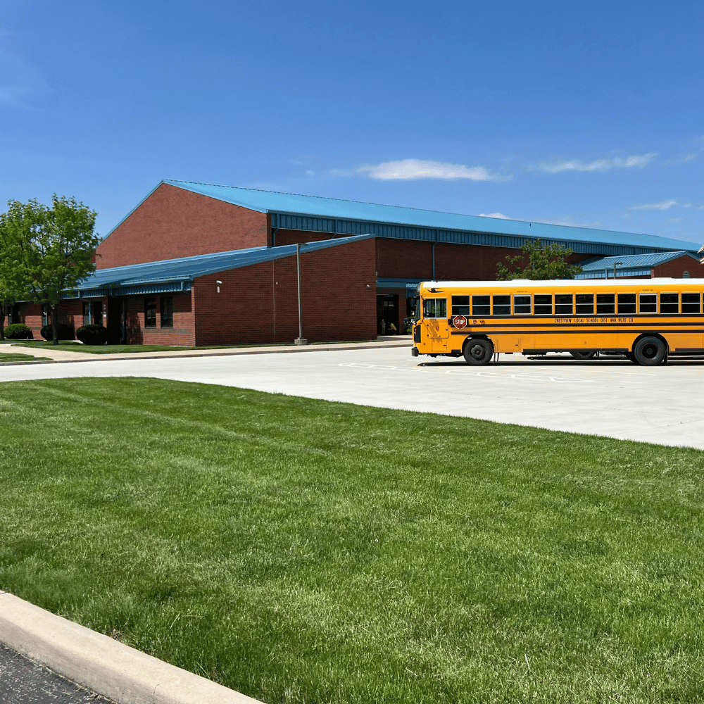 A yellow school bus parked outside a red brick school building with a blue roof, under a clear blue sky.