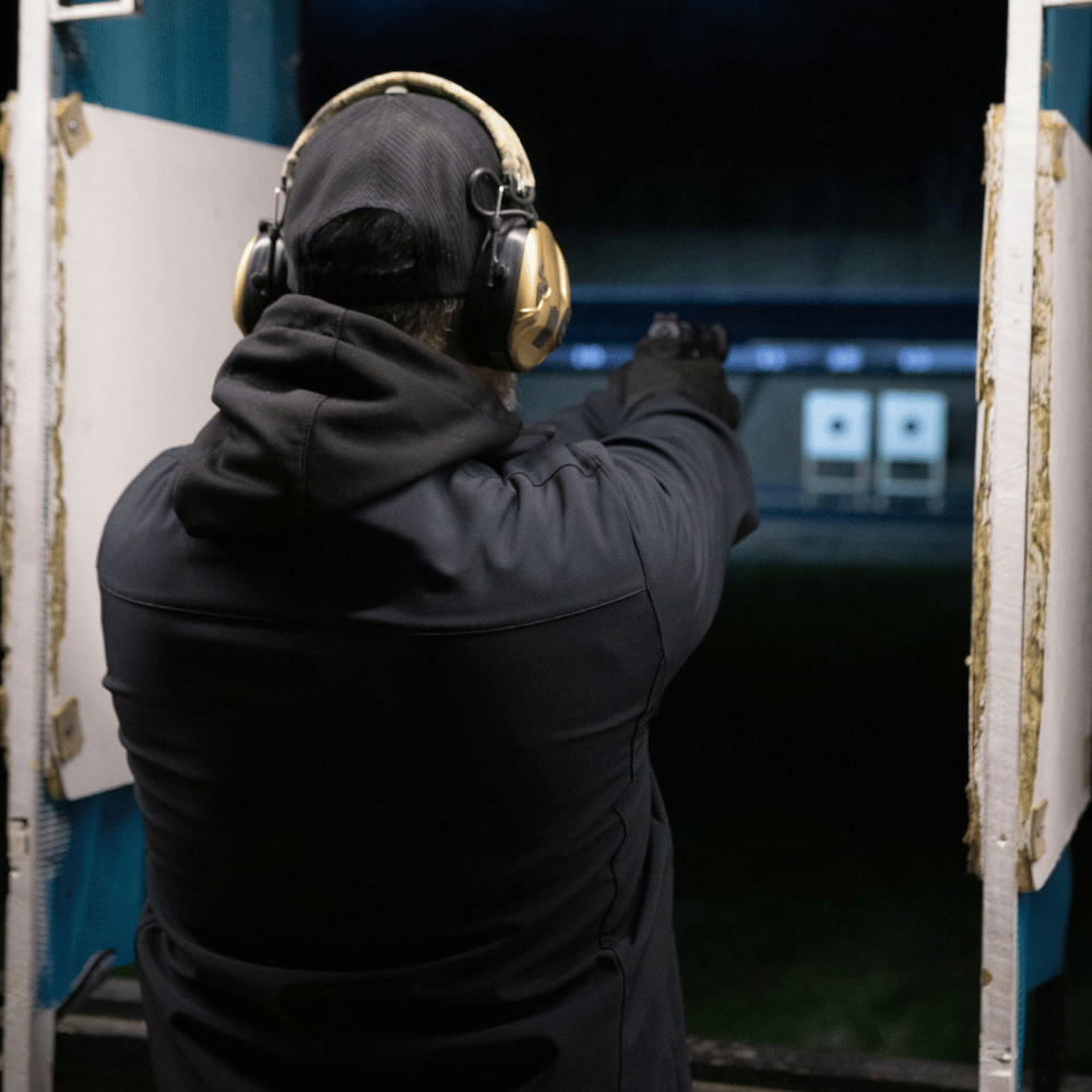A person in dark clothing and a black beanie, wearing hearing protection, aiming a handgun at an indoor shooting range with targets in the distance. The number '17' is displayed above the shooting area.