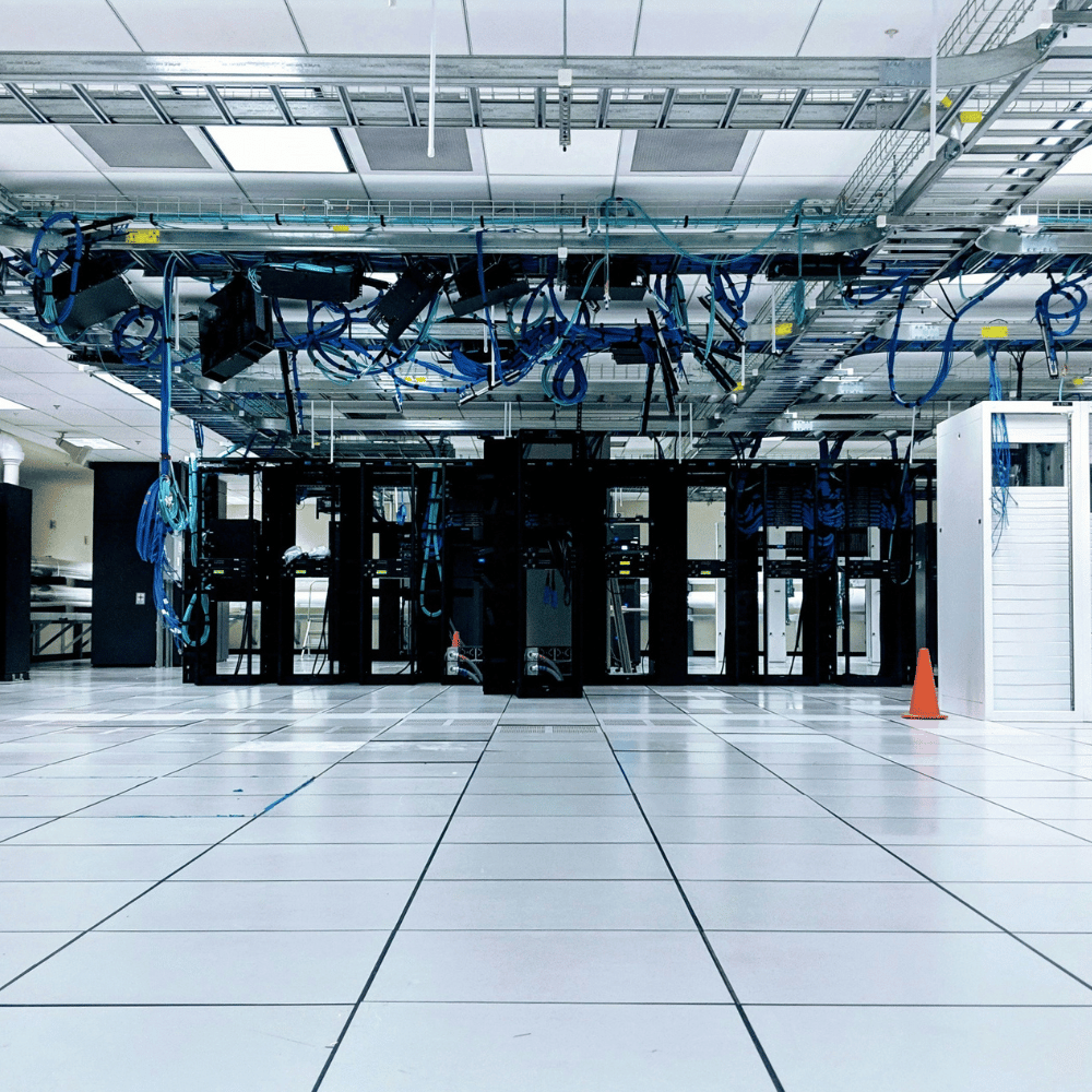 Empty server room with black server racks, blue cables hanging from the ceiling, a white cabinet, and a person working at a desk in the background.