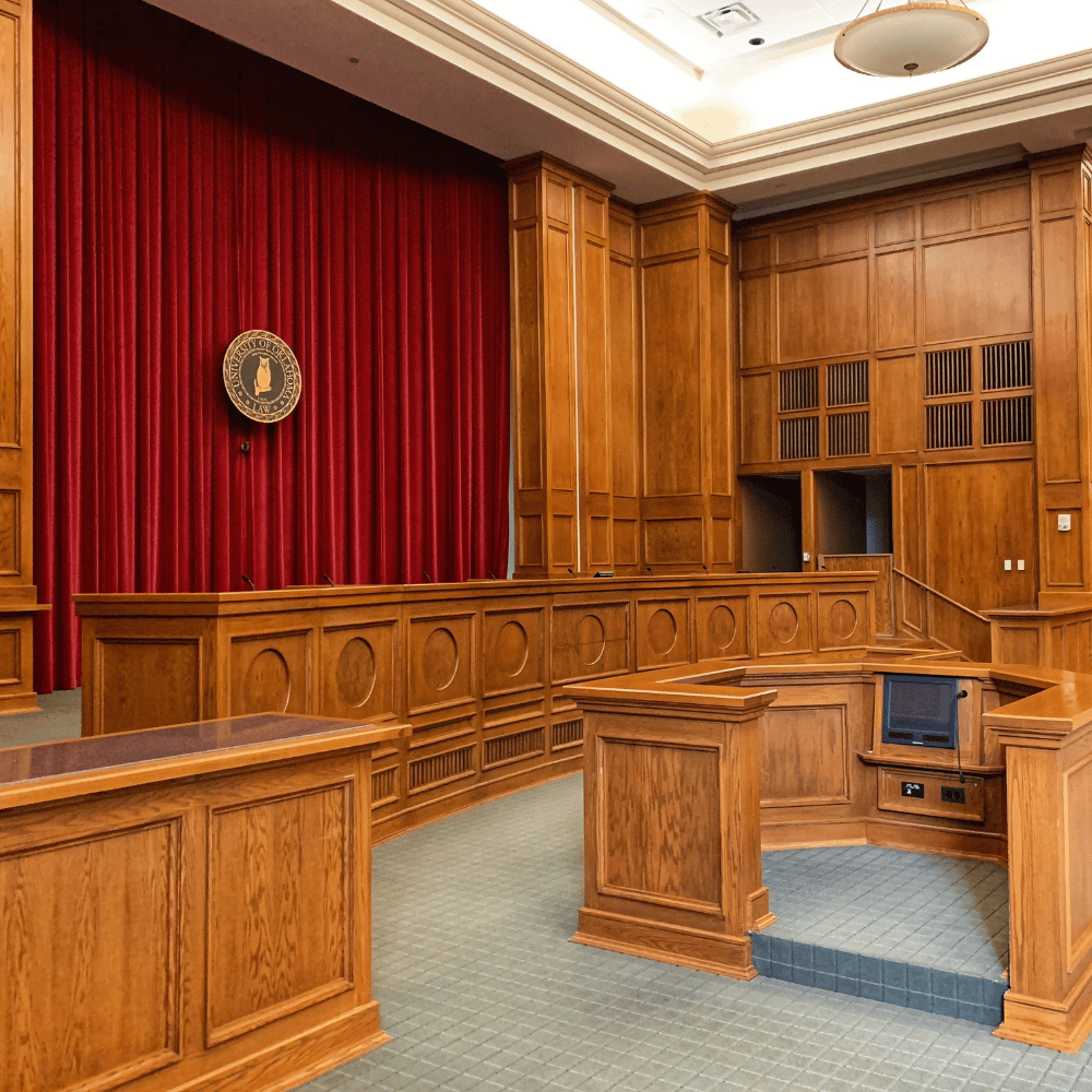 Interior of a courtroom with wooden paneling, a red curtain, a podium, and a framed painting of a person in traditional clothing.