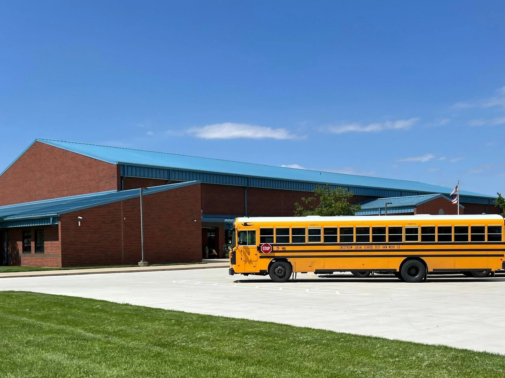 A yellow school bus parked outside a red brick school building with a blue roof, under a clear blue sky.