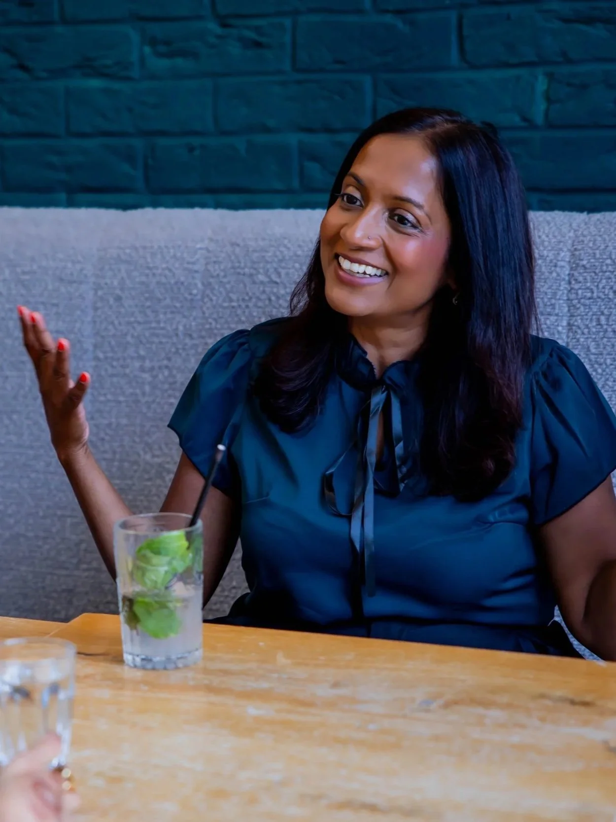 Smiling woman with dark hair and dark blue top sitting at a table with a mint soda drink in a glass in front of her, gesturing with her right hand.