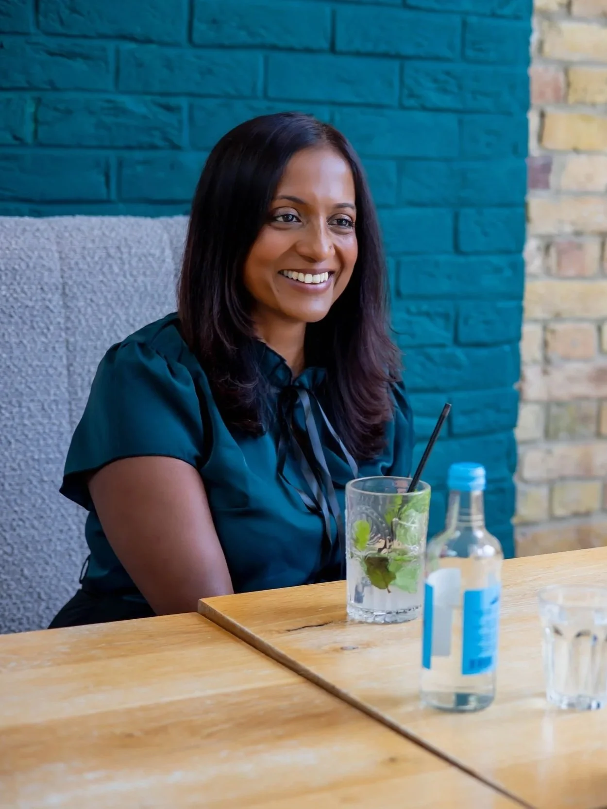 A woman sitting at a wooden table, smiling, with a glass of water with mint leaves, a small bottle of hand sanitizer, and a blue-capped water bottle.