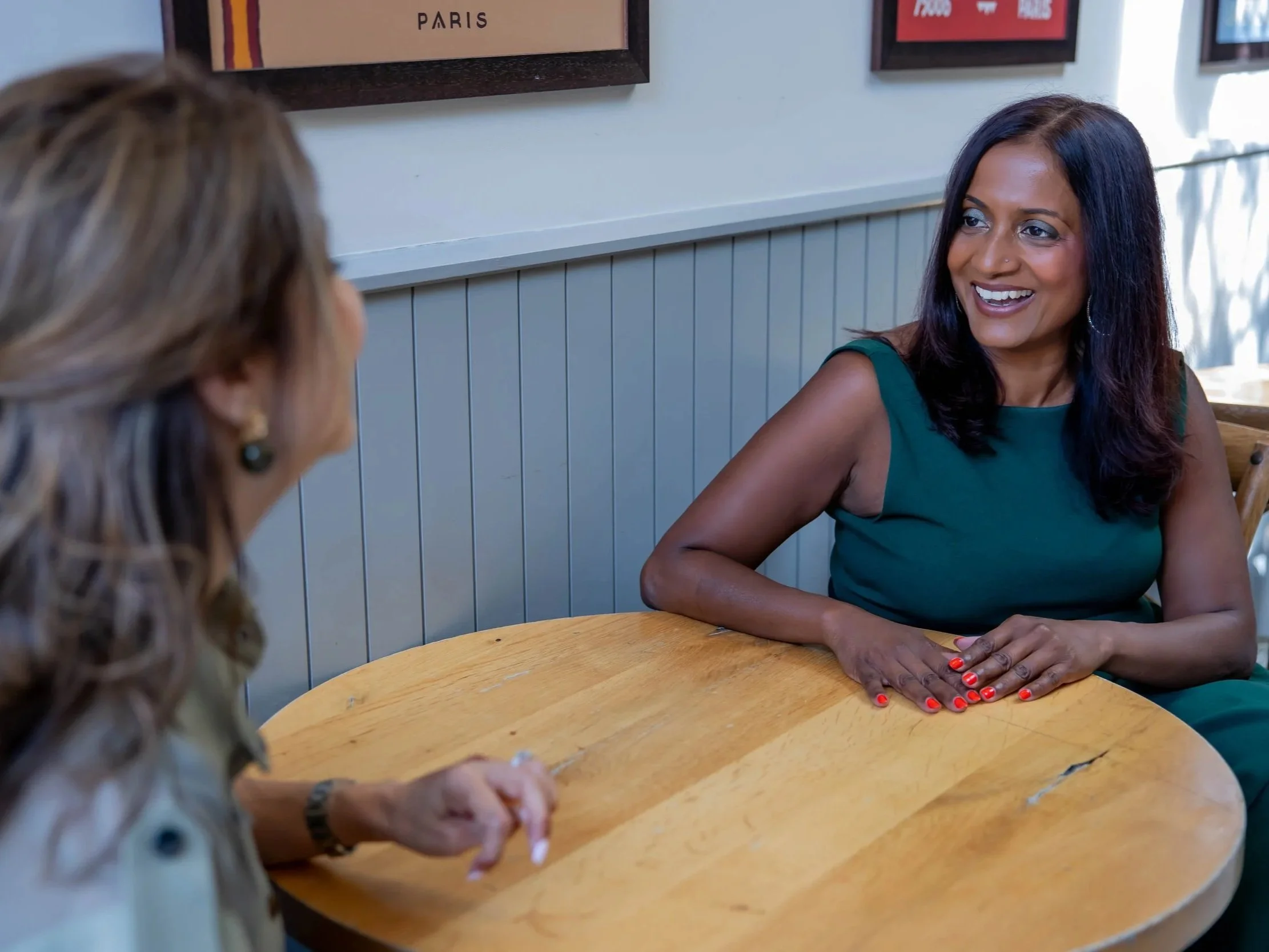 Two women sitting at a round wooden table inside a restaurant or cafe, engaged in conversation. The woman in the foreground has long, dark hair, blue eyeshadow, a nose piercing, and is wearing a green sleeveless top with bright red nail polish. The woman in the background has curly hair, earrings, and is wearing a patterned blouse. The background includes a white wall with framed artwork and beadboard paneling.