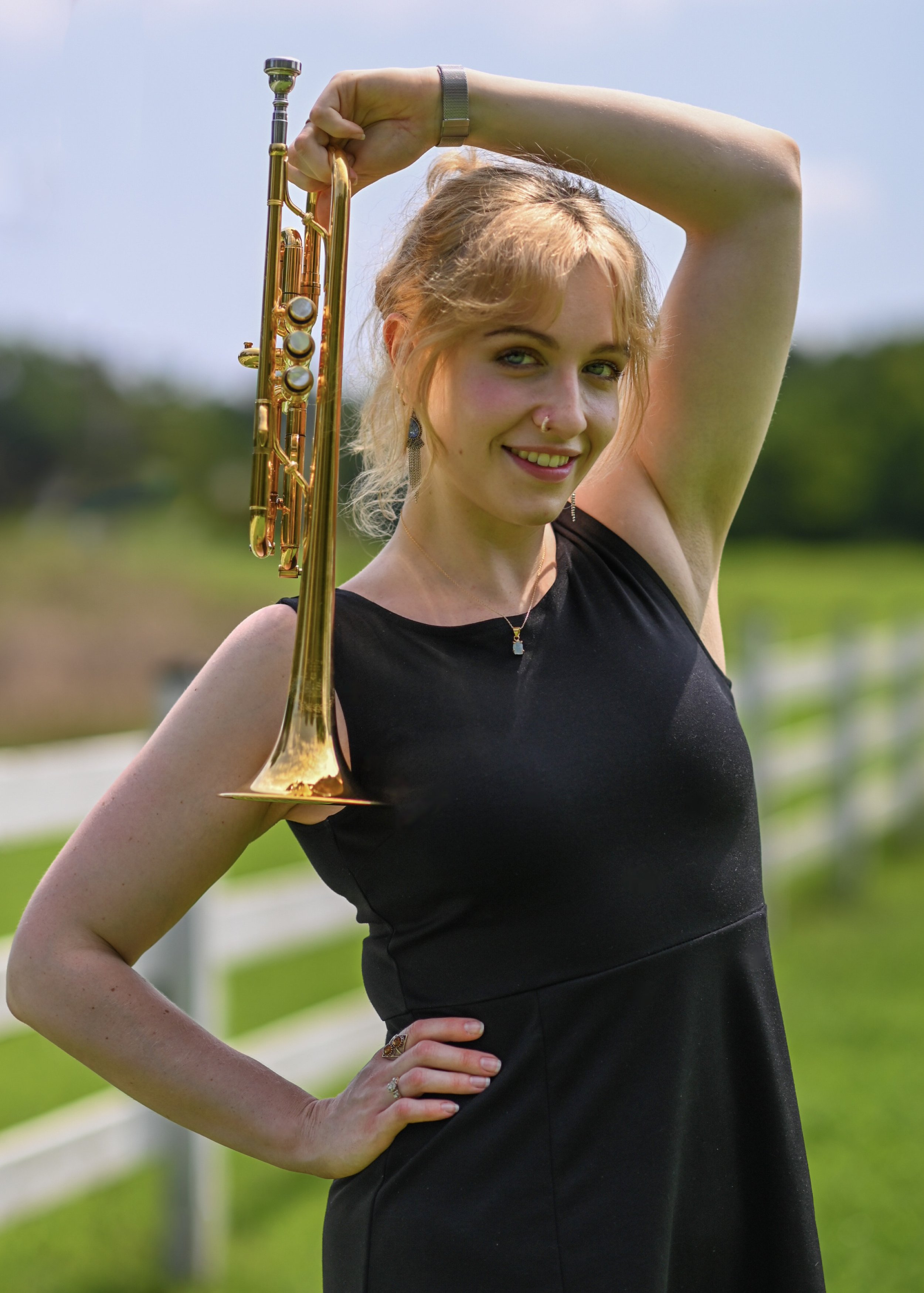 A woman with blonde hair, wearing a black dress and silver jewelry, holding a gold trumpet over her shoulder outdoors with a green field and blue sky in the background.