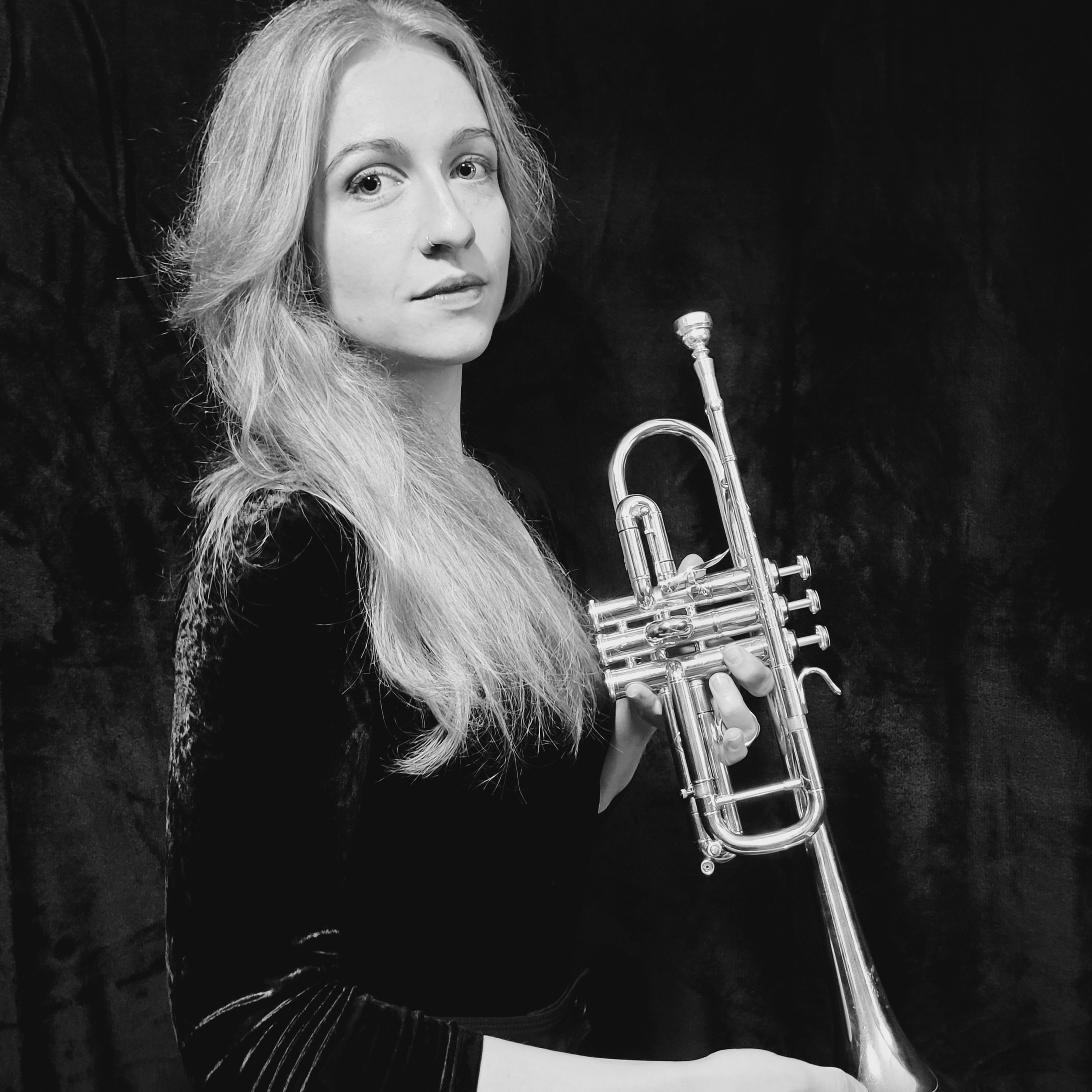 A black-and-white photo of a young woman with long hair holding a trumpet. She has a nose ring and is looking at the camera with a serious expression against a dark background.