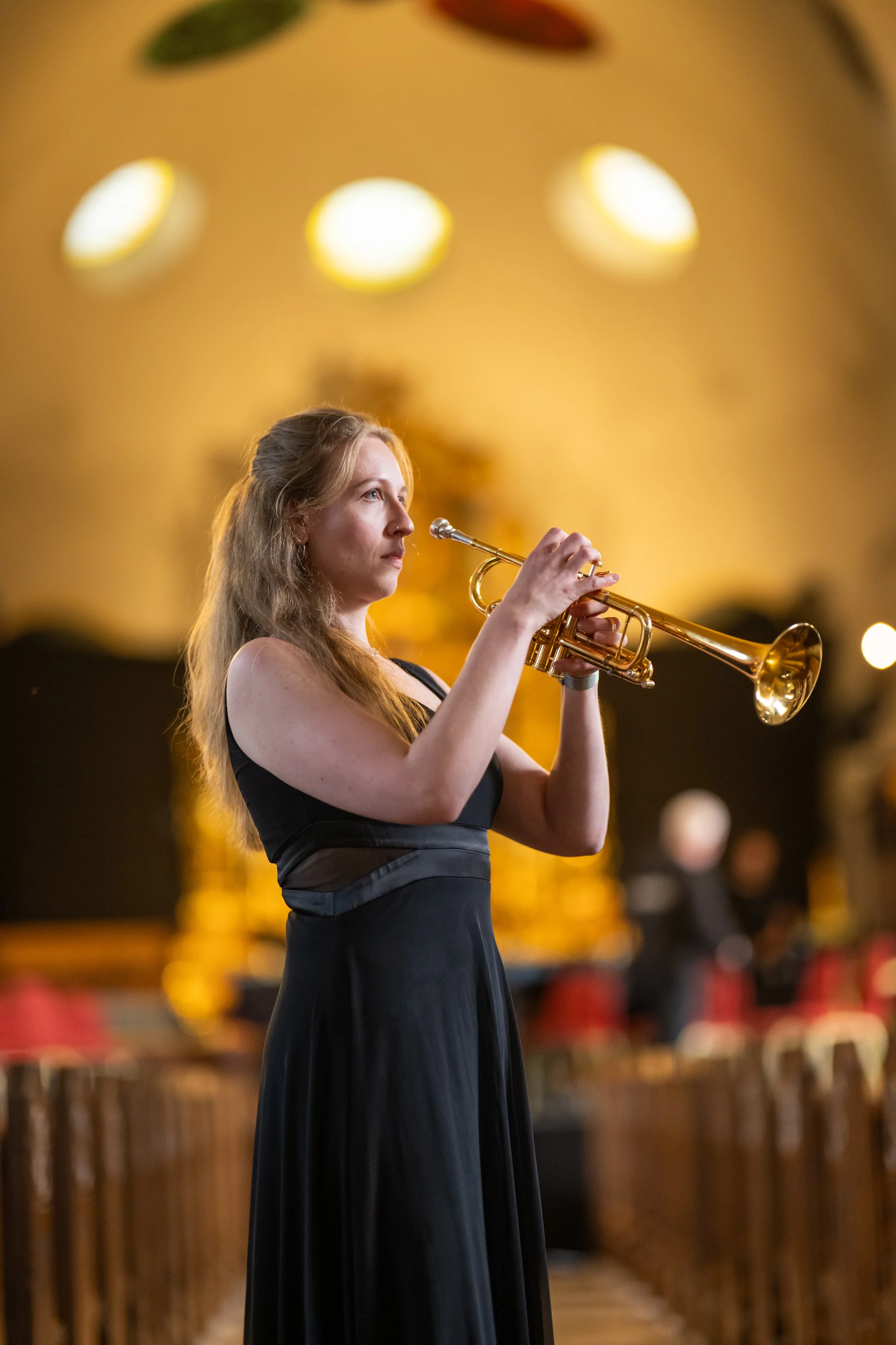 A woman in a black dress playing a trumpet indoors with blurred background and warm lighting.