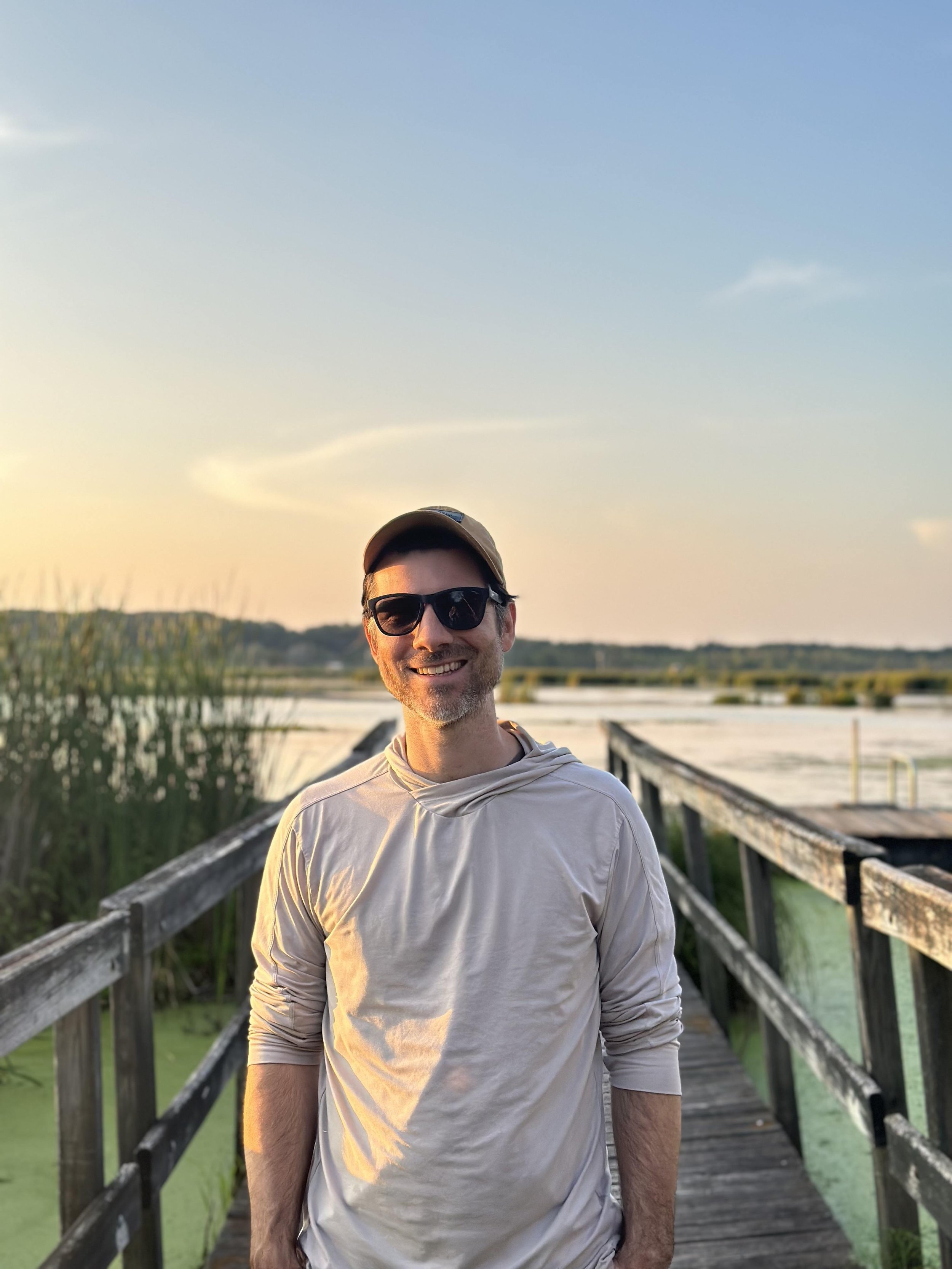 A man wearing sunglasses, a cap, and a light-colored long-sleeve shirt standing on a wooden bridge over water during sunset or sunrise. There are trees and a cloudy sky in the background.
