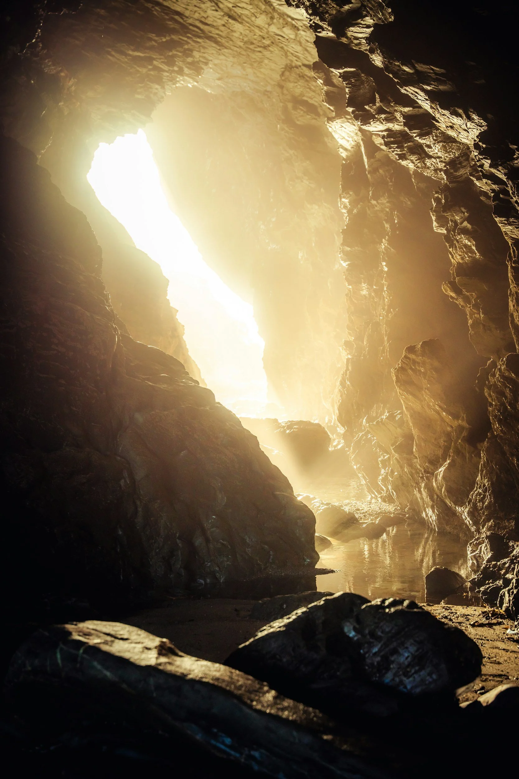 Sunlight streaming into a dark cave, illuminating the rocky walls and reflecting off a pool of water at the cave's entrance.