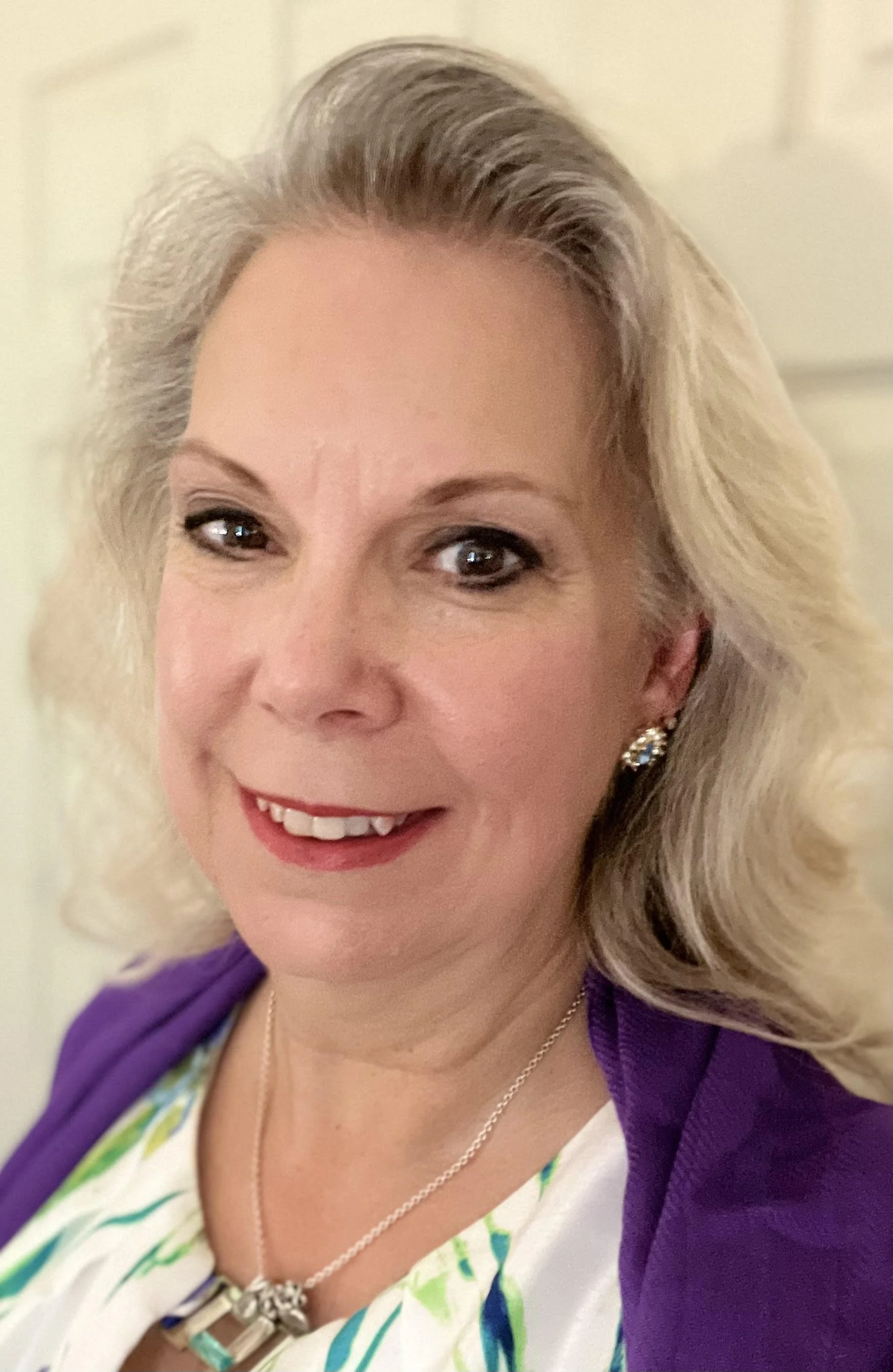 Close-up photograph of a smiling woman with platinum blonde hair styled in waves. She is wearing earrings, a necklace with a pendant, and a purple blazer over a patterned top.
