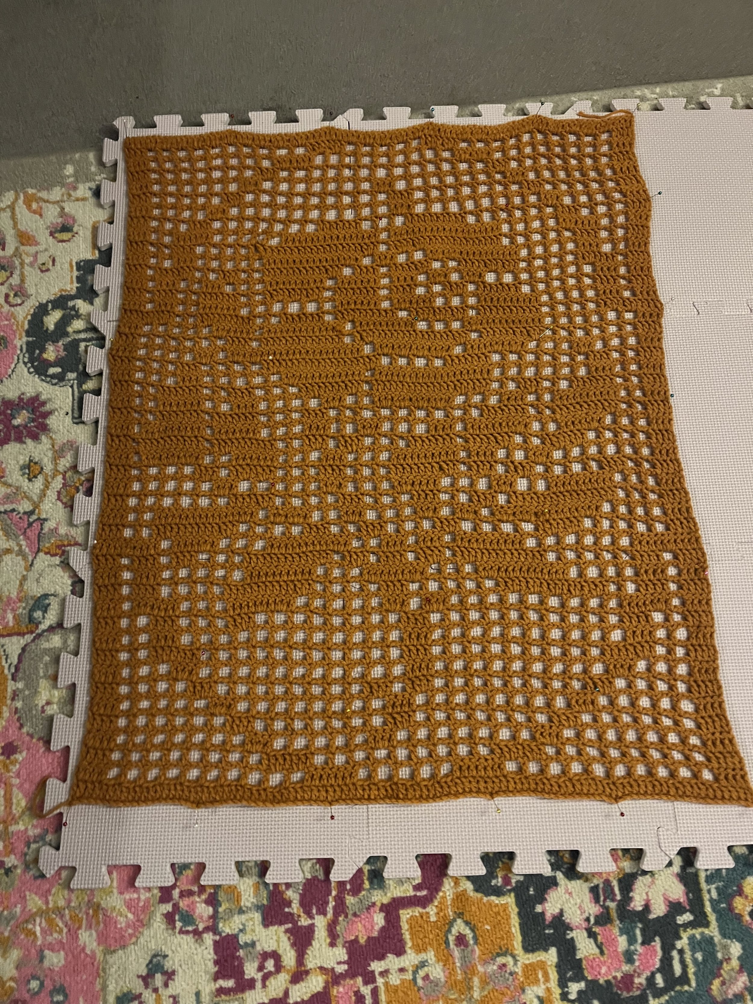 A crocheted orange square with a floral pattern in the middle, placed on a foam puzzle mat on top of a colorful patterned carpet.