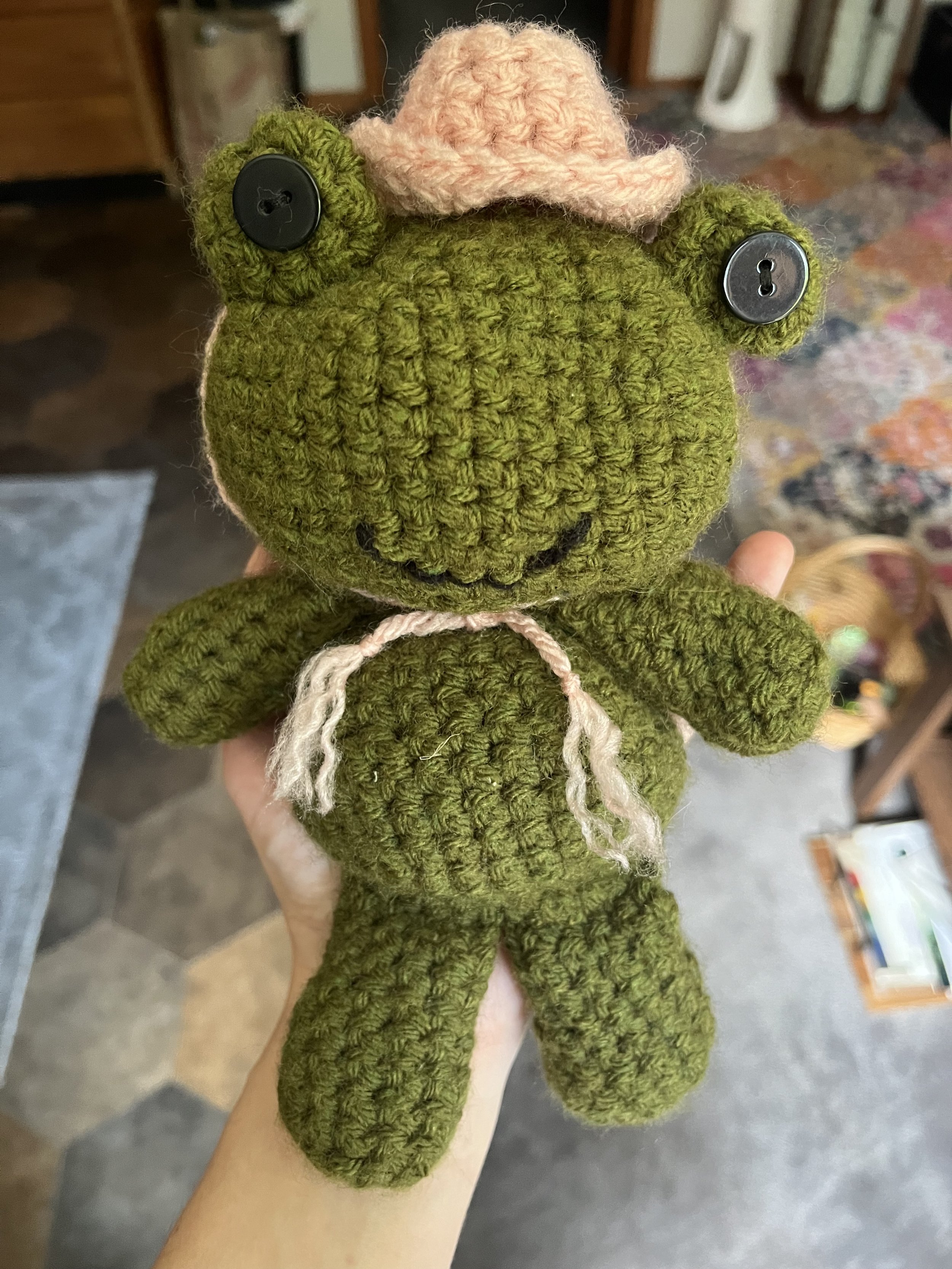 A crocheted stuffed frog with a pink hat and string around its neck, held by a person's hand.
