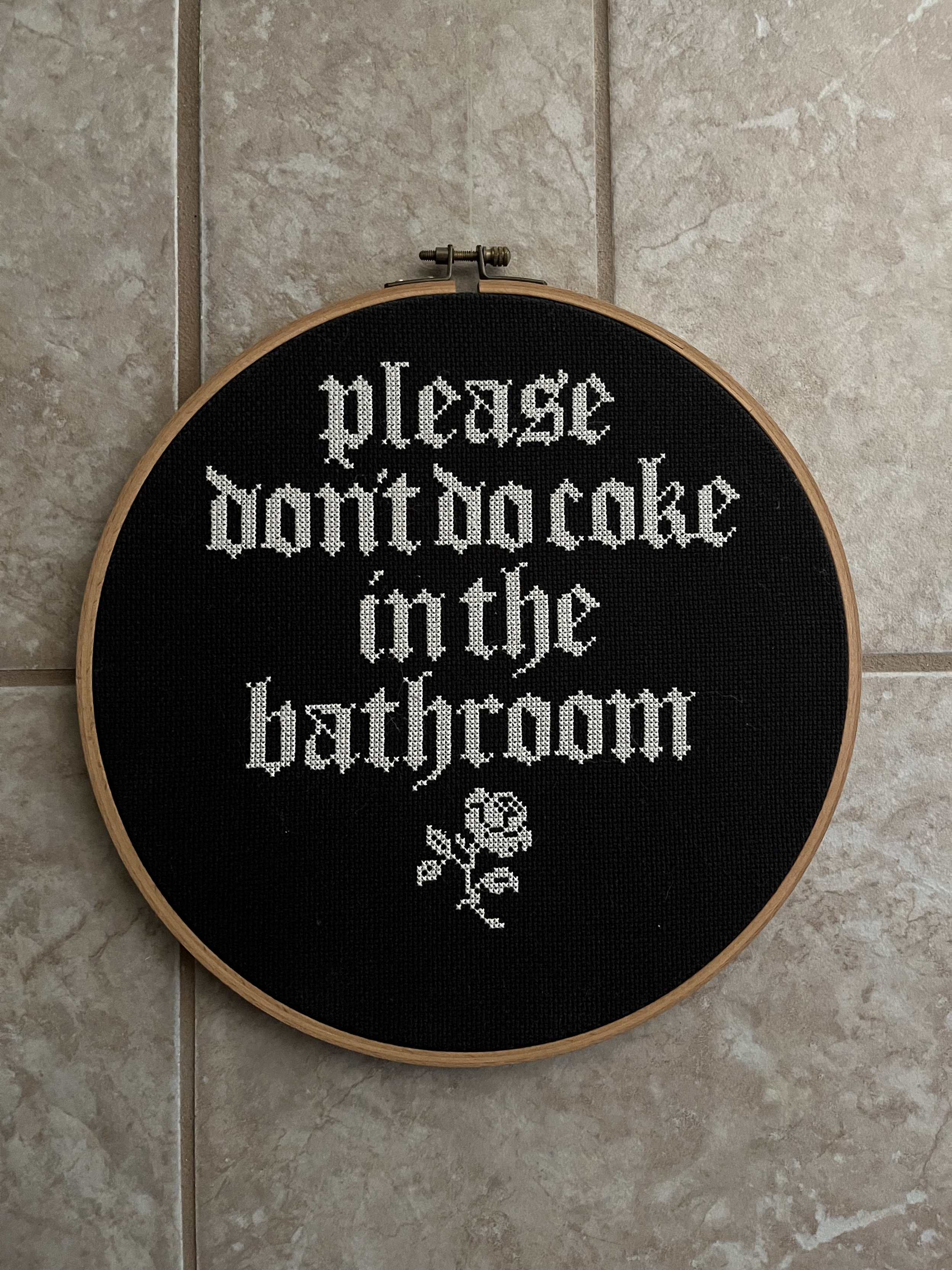 A black circular embroidery hoop with a cross-stitched message that reads "please don't do coke in the bathroom" and a small decorative flower pattern below the text, hanging on a tiled wall.