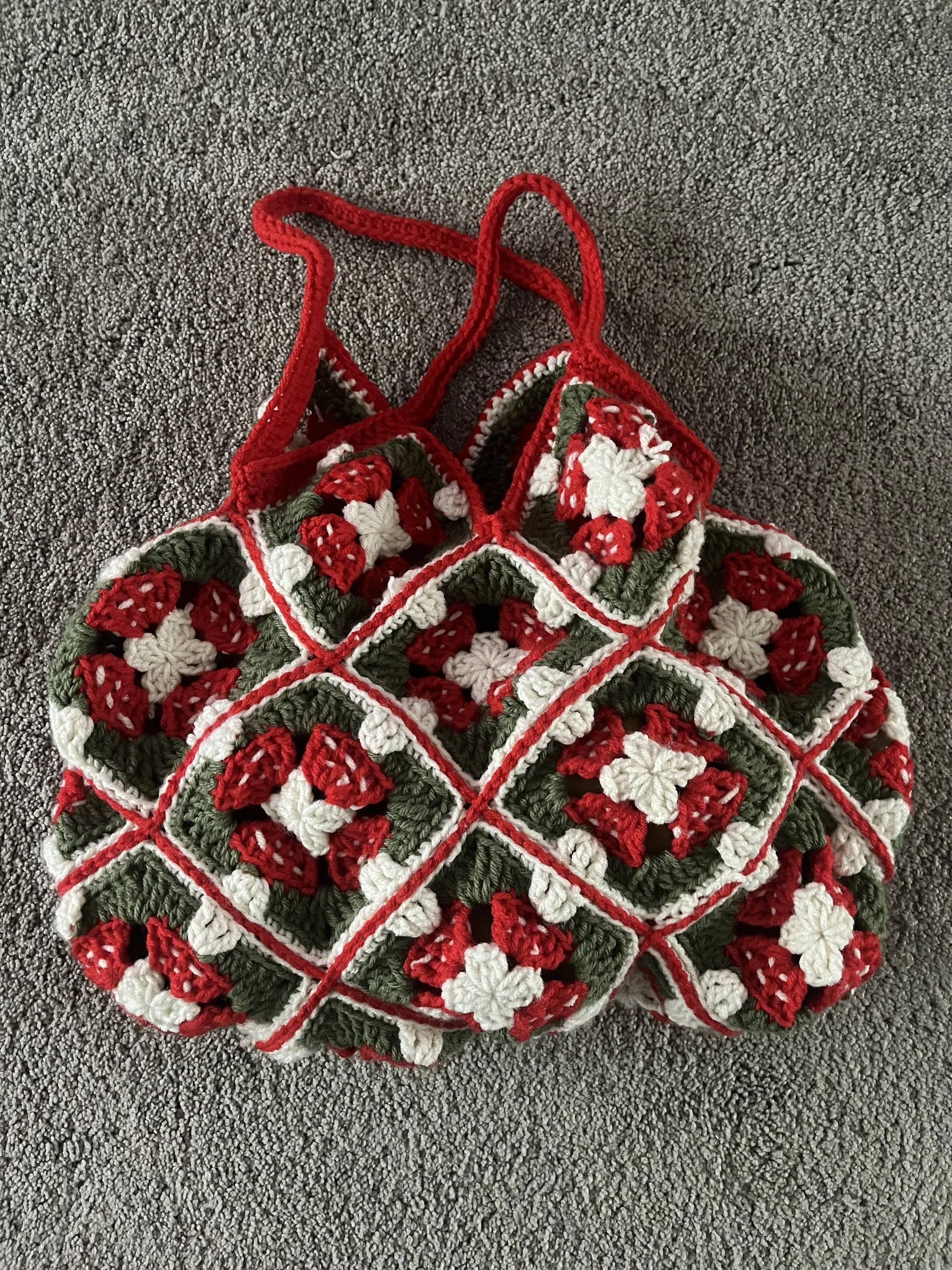 A crocheted bag with a mushroom pattern of red, white, and green colors, lying on a grey textured carpet.