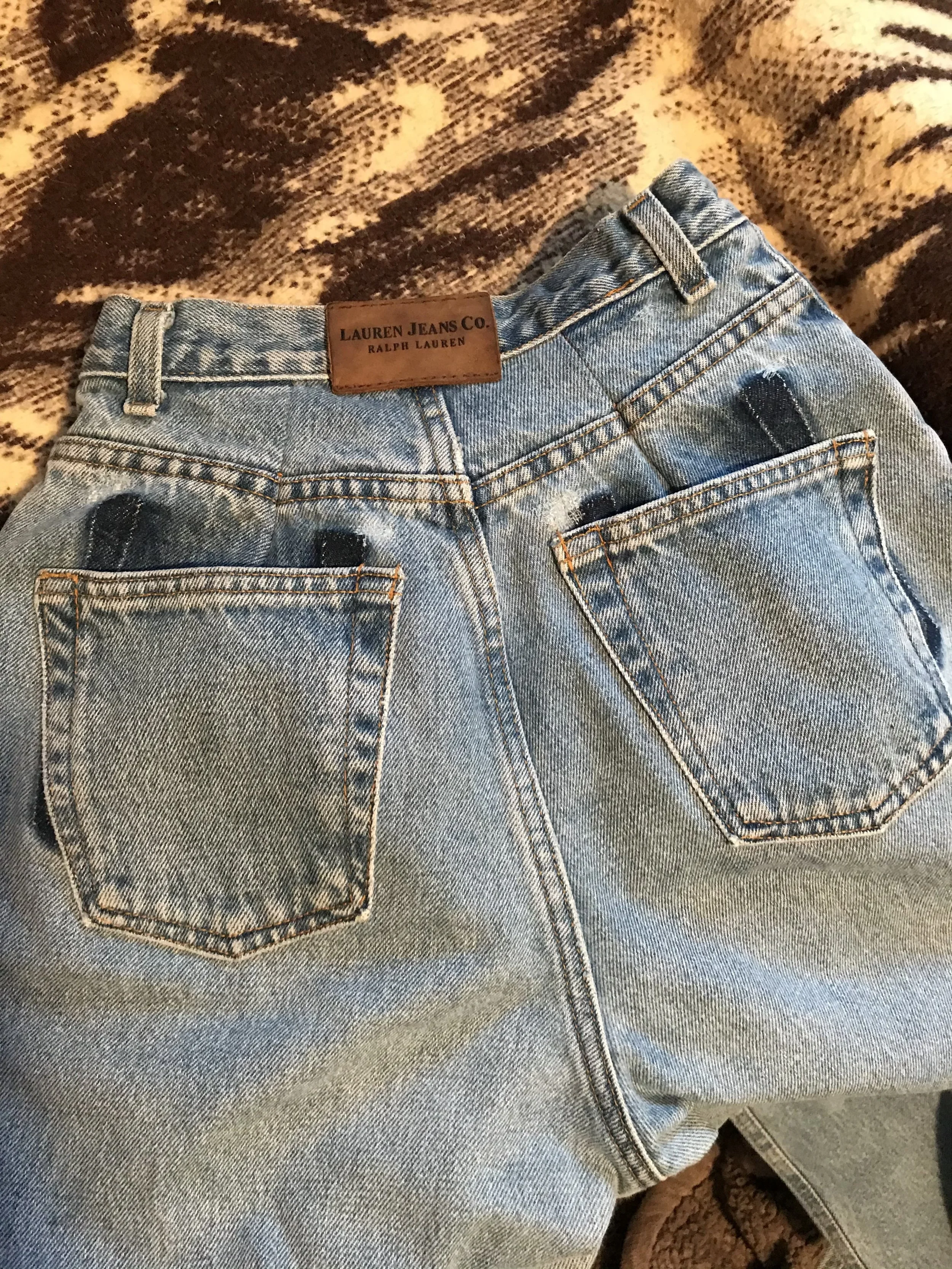 Pair of blue denim jeans with a brown leather patch labeled 'LAUREN JEANS CO. RALPH LAUREN' on the waist, lying on a patterned blanket.