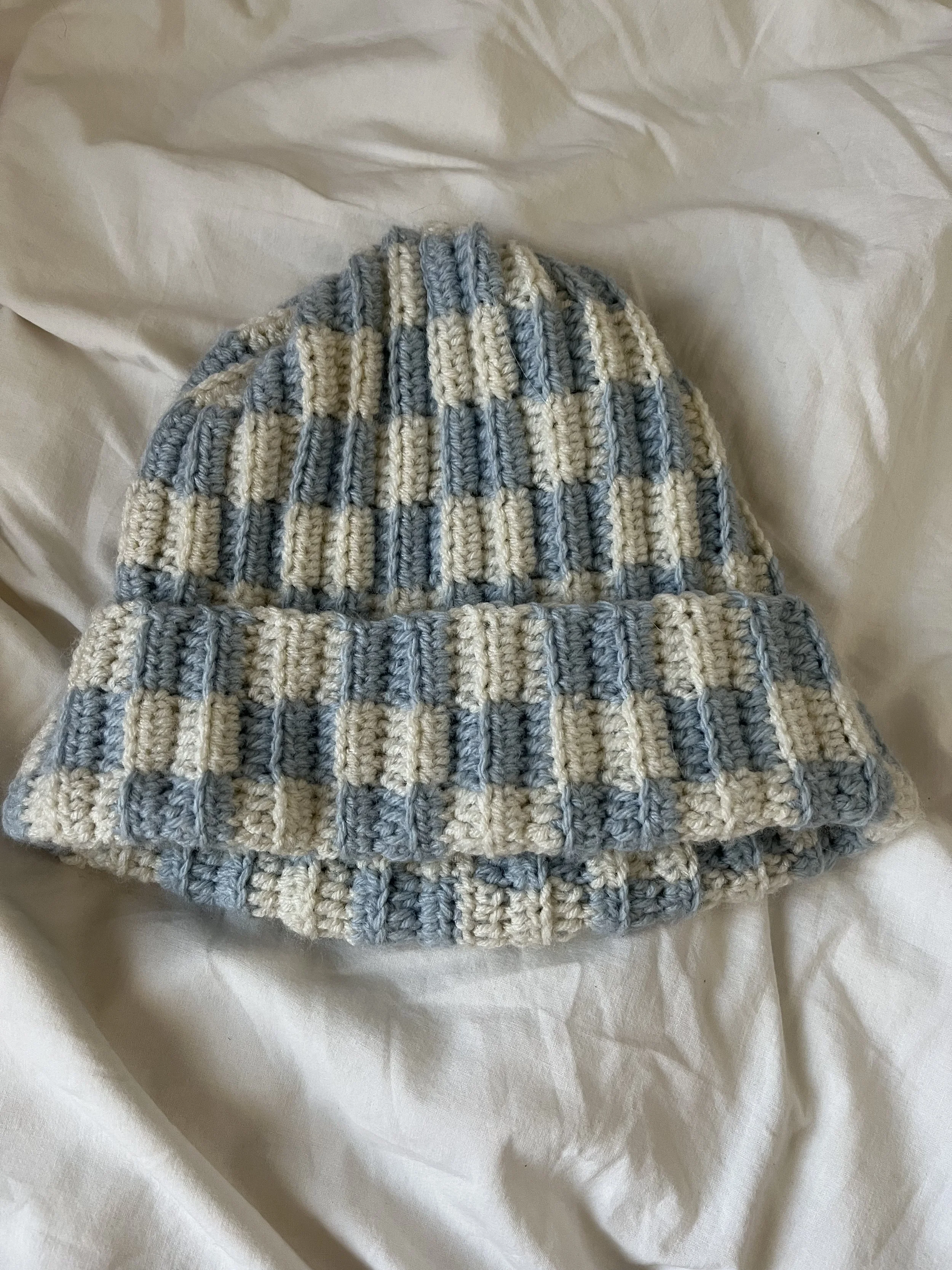 A crocheted beanie hat with a checkered pattern of blue and white stripes, resting on white fabric.