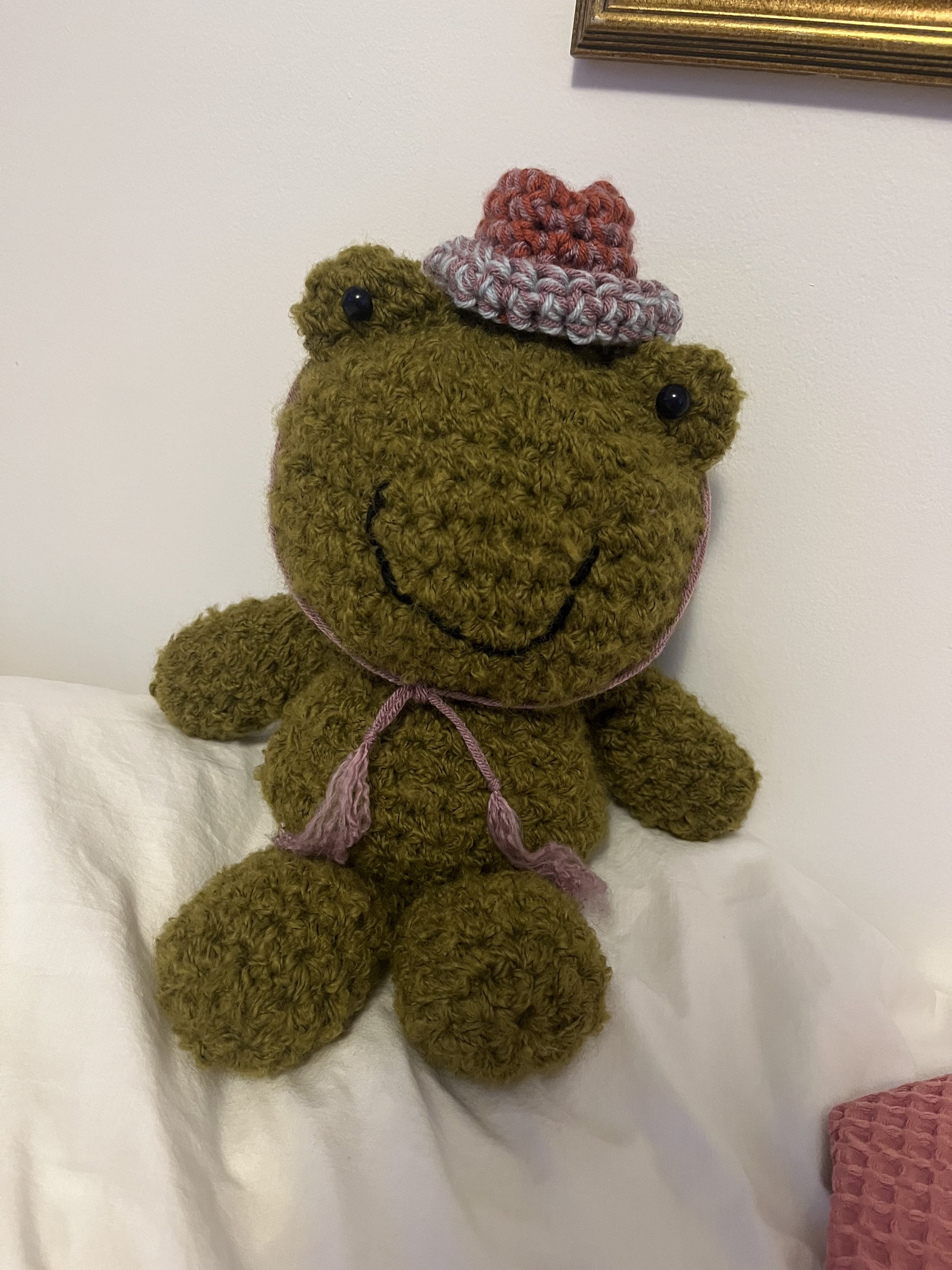 A stuffed frog with a pink yarn bow around its neck, wearing a small pink and gray crocheted hat, sitting against a white wall.