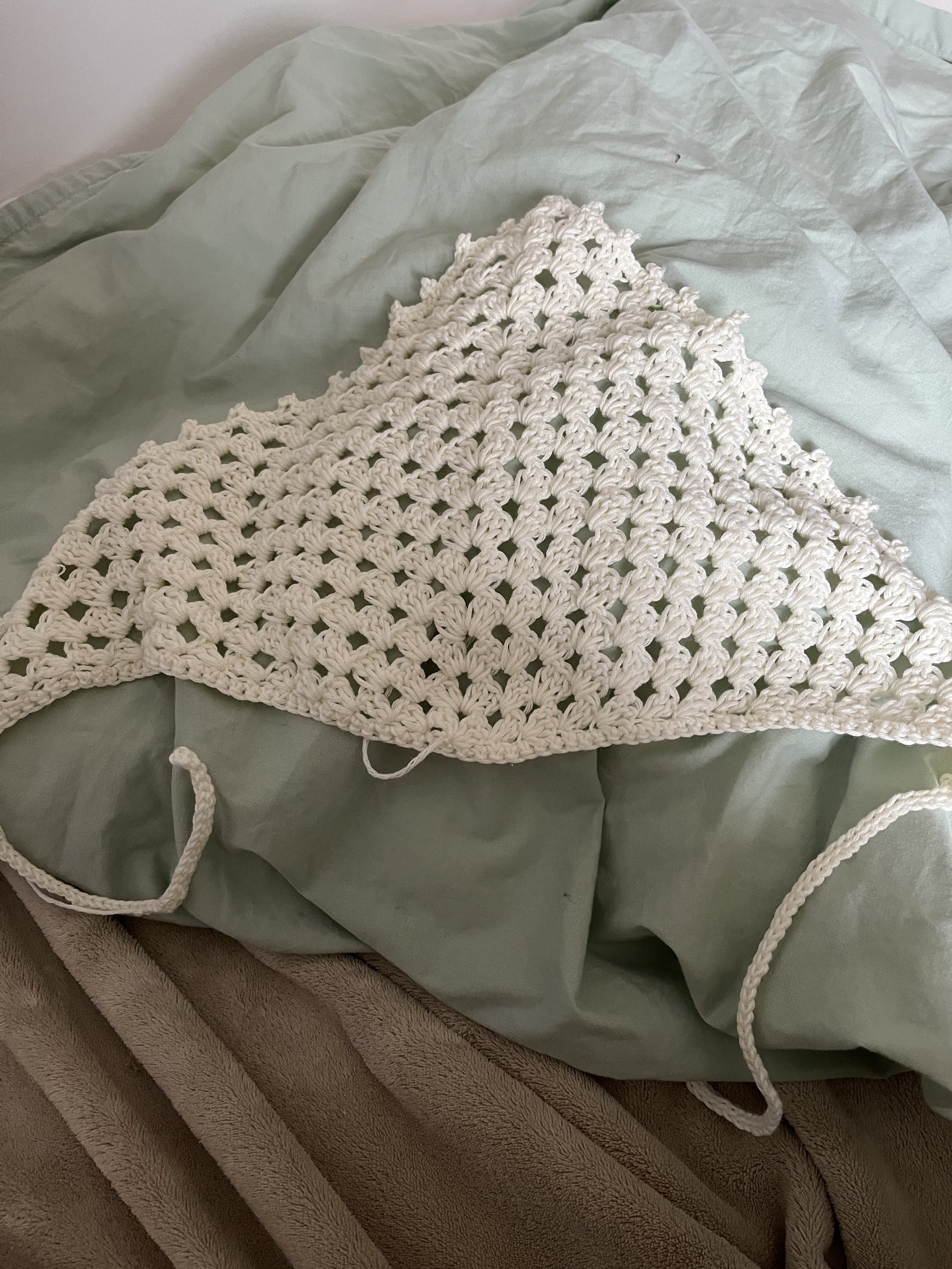 A white crochet bandana resting on a light green blanket, which is draped over a brown blanket.