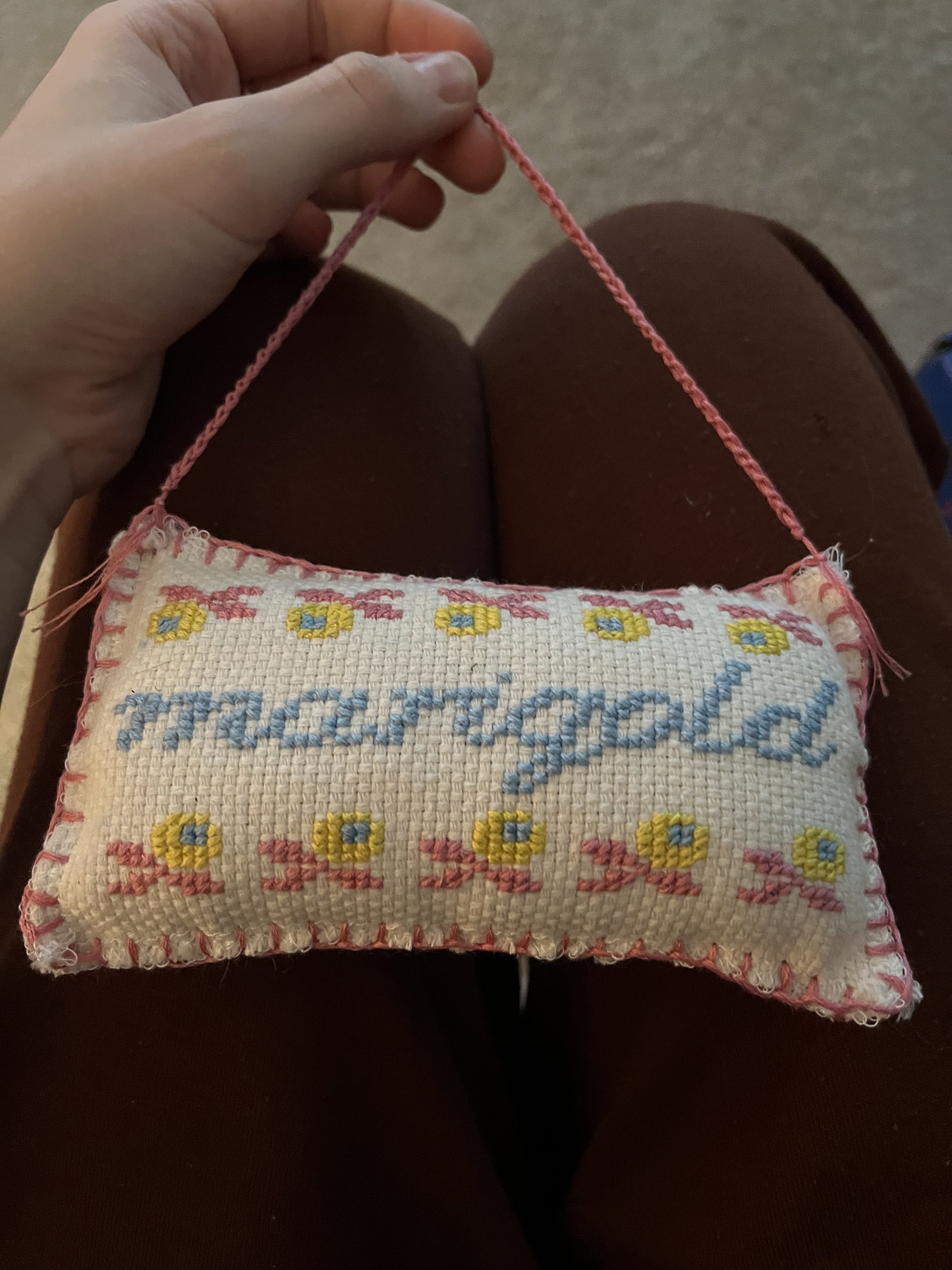 A person's hand holding pink embroidery floss with a piece of cross-stitched fabric showing a pattern of flowers and the word 'marigold' in blue.