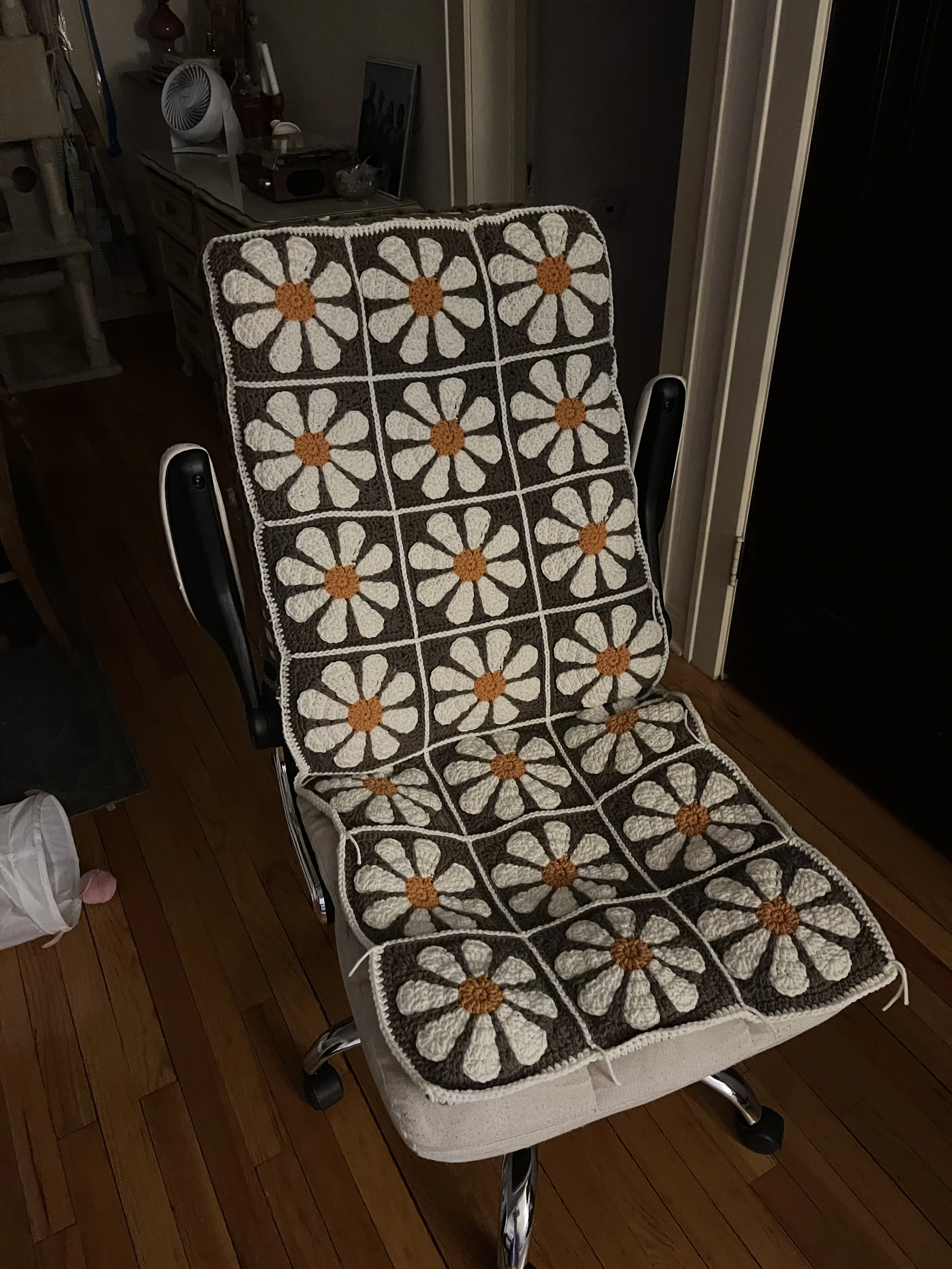 A desk chair with a crochet covering featuring a floral pattern in white, brown, and orange on the seat and backrest, located in a room with wooden flooring.