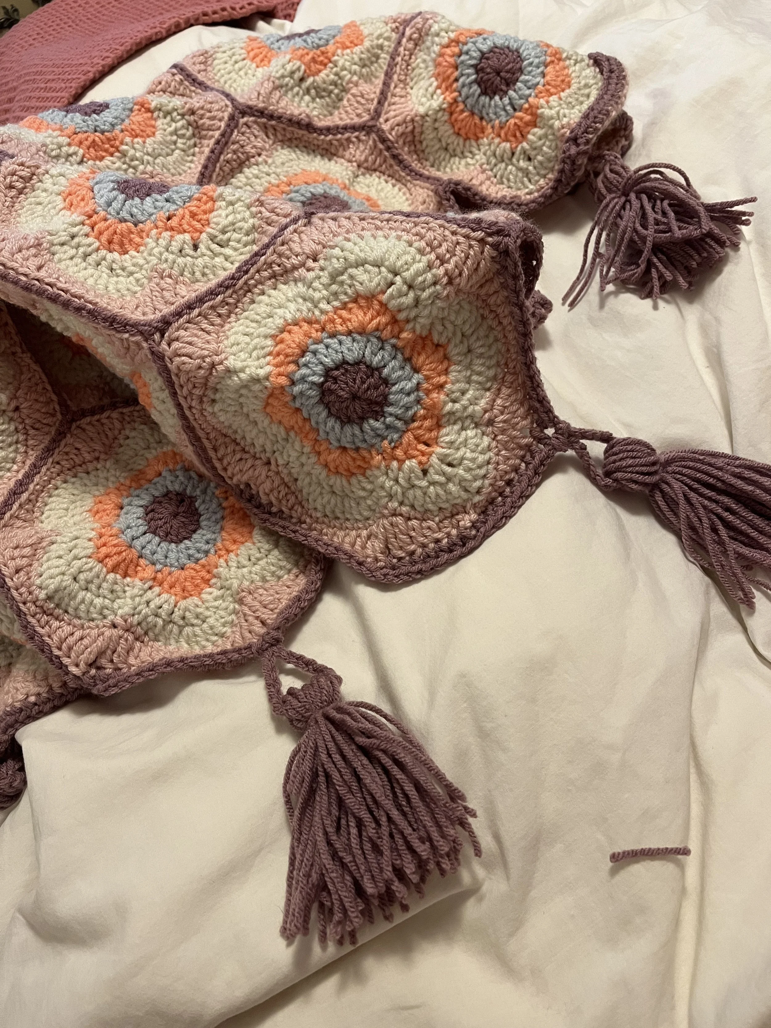 A crochet blanket with a colorful circular pattern in shades of purple, orange, gray, and cream, draped on a white bed with tassel edges.