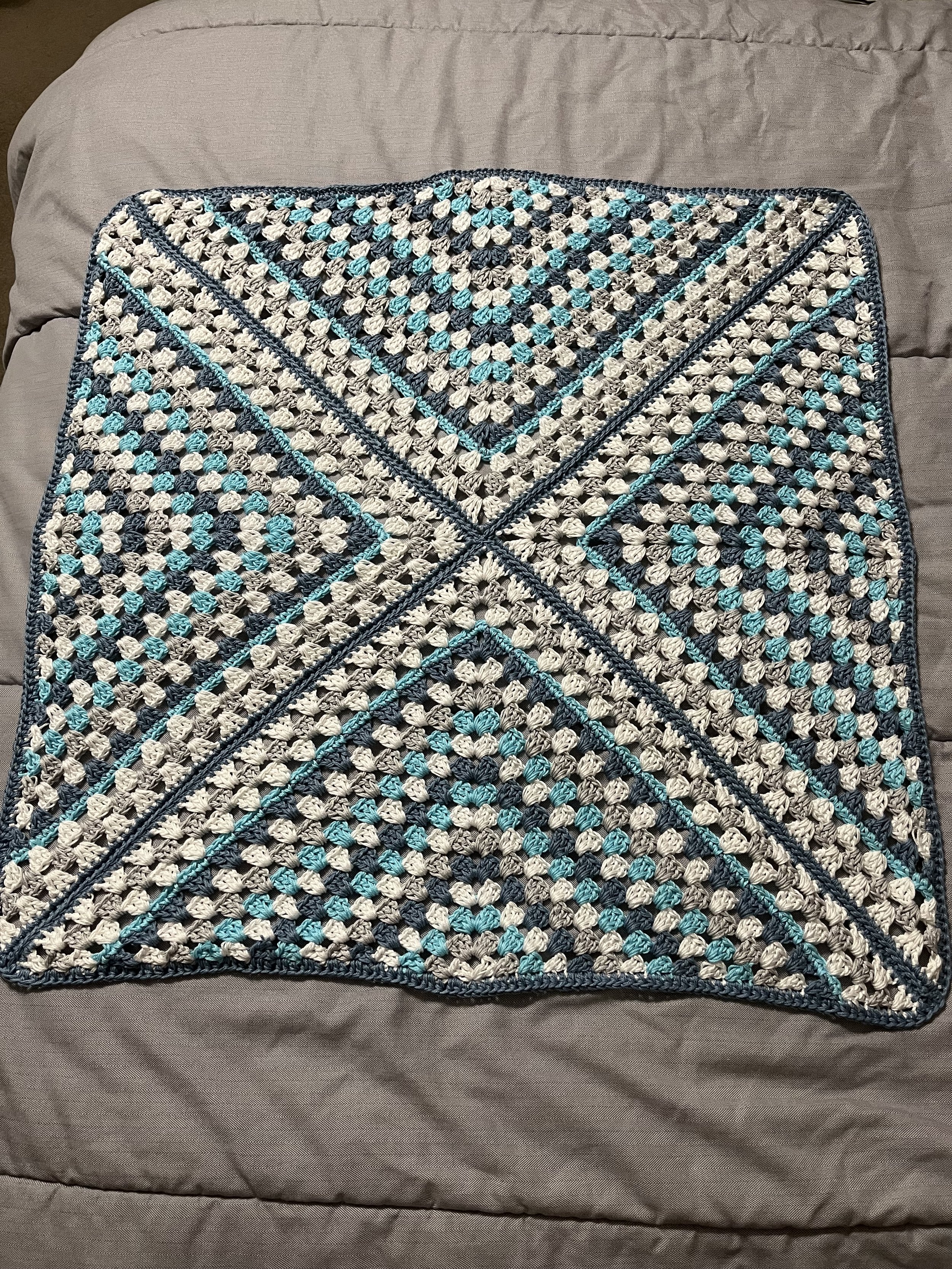 A layered crochet blanket with diagonals and geometric patterns in shades of blue, gray, white, and beige, placed on a bed with a beige quilted bedspread.