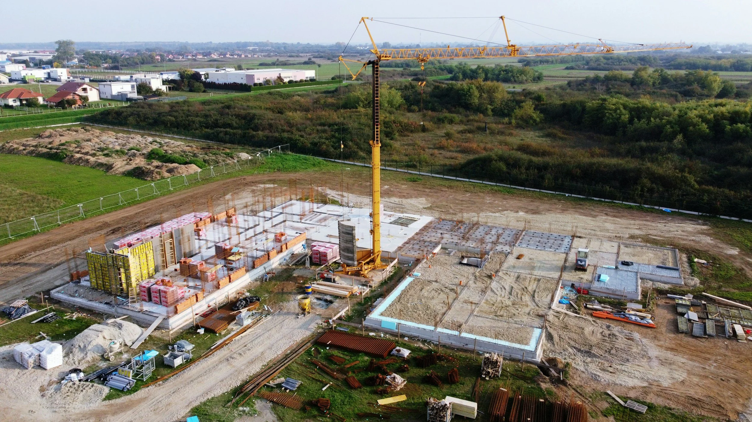 Une construction en cours avec une grue jaune, des fondations et des structures en béton, entourée d'un terrain partiellement aménagé, avec des maisons et des zones vertes à l'arrière-plan.